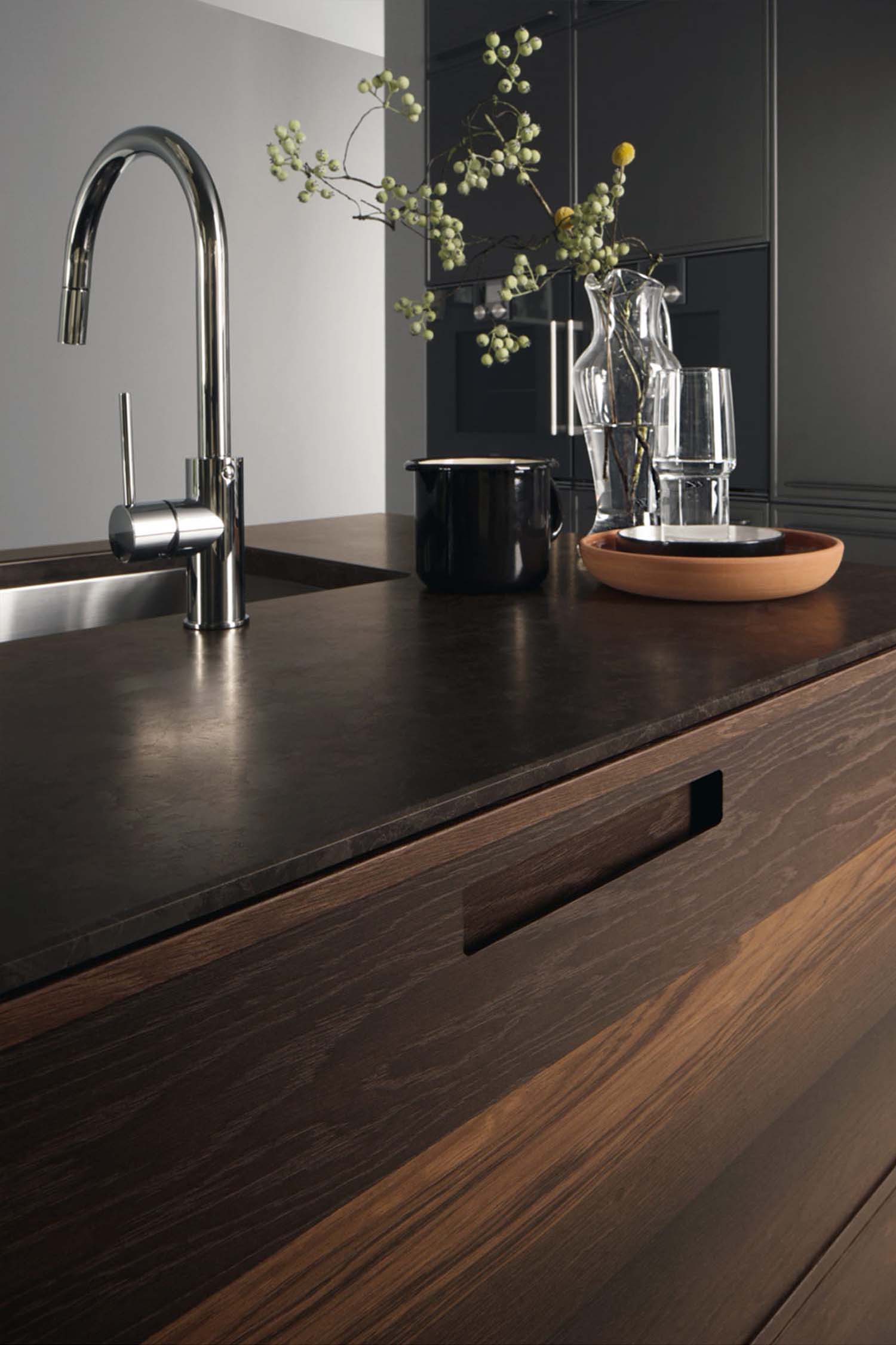 A featured image of a luxury industrial kitchen with a large, dark kitchen island that has a hob and seating. The back wall features light, concrete-effect tall cabinets and a wooden slatted panel. The space has a high ceiling with exposed pipes, an industrial-style extractor fan above the island, and large windows that fill the room with natural light.