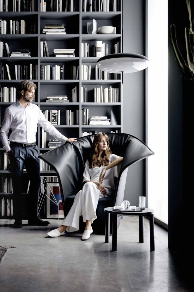 A modern living space with a black leather wingback armchair and a man and a woman. The woman is seated in the chair, wearing white pants and a white shirt. The man stands to the left of the chair, leaning on its high back, wearing a white button-down shirt and dark pants. The armchair has a unique fan-shaped, pleated back. A small black side table with a glass and a book on it is next to the chair. In the background, a large, floor-to-ceiling bookshelf is filled with books and decorative objects.