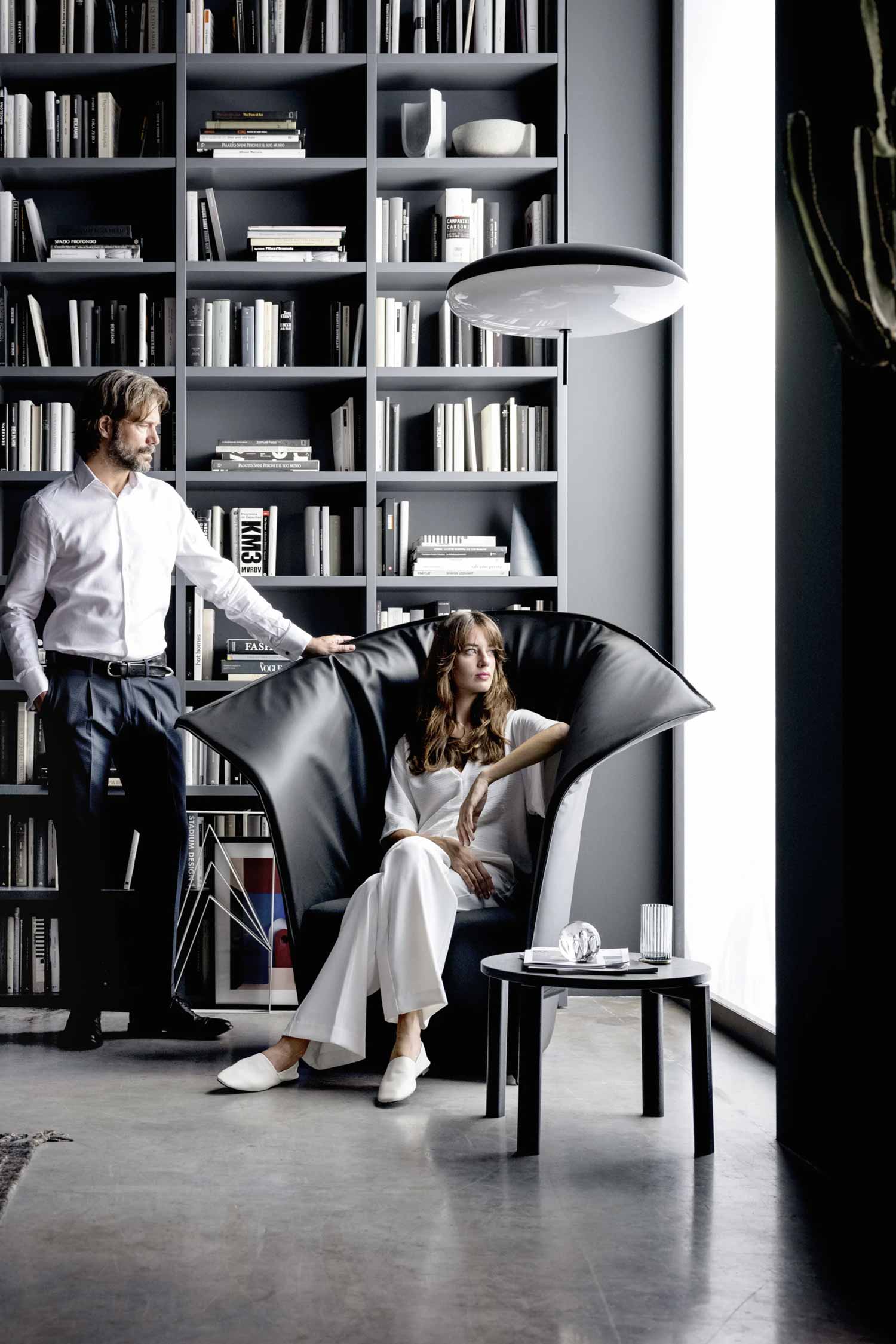 A modern living space with a black leather wingback armchair and a man and a woman. The woman is seated in the chair, wearing white pants and a white shirt. The man stands to the left of the chair, leaning on its high back, wearing a white button-down shirt and dark pants. The armchair has a unique fan-shaped, pleated back. A small black side table with a glass and a book on it is next to the chair. In the background, a large, floor-to-ceiling bookshelf is filled with books and decorative objects.