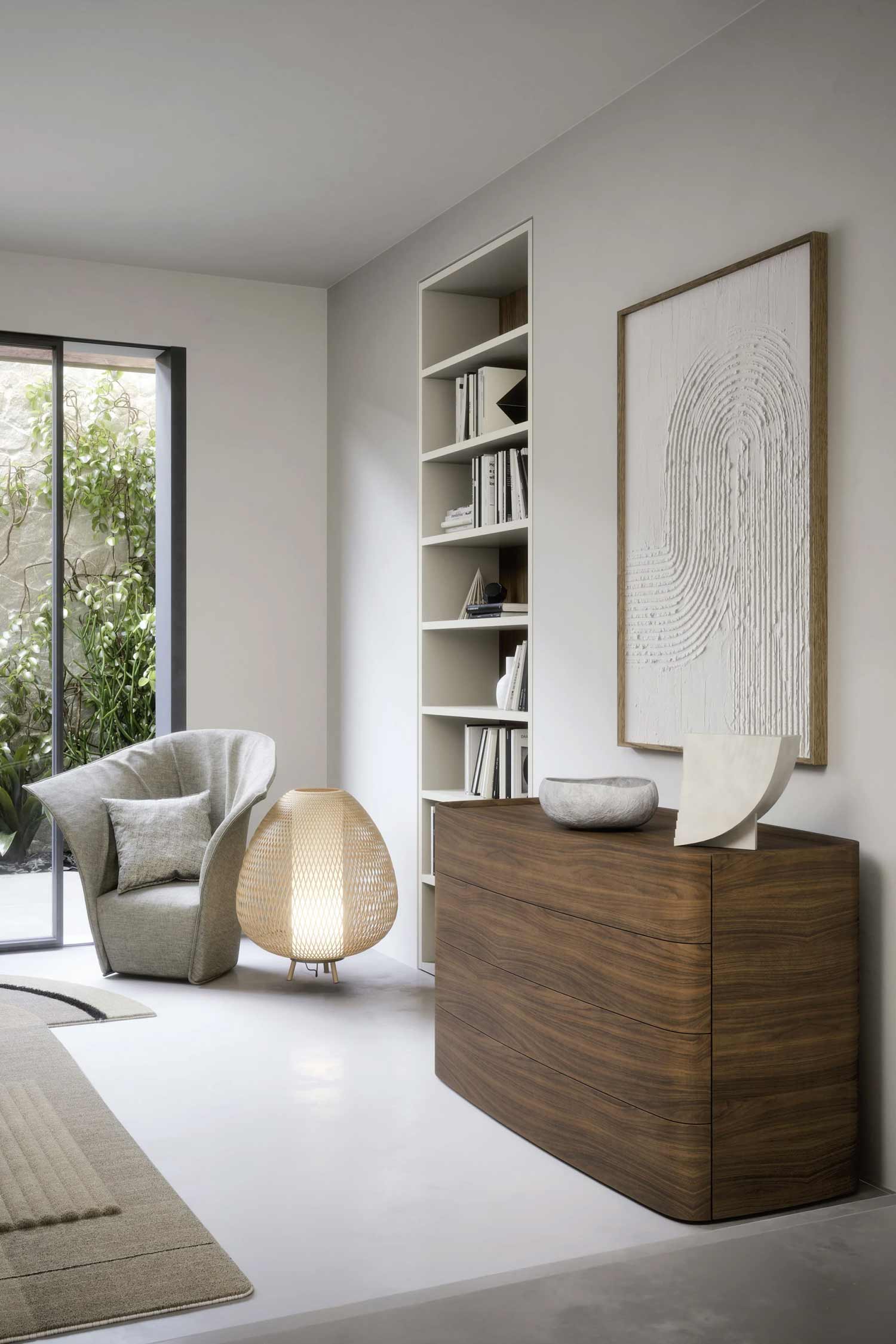 A large, light-filled room features a modern wood and white chest of drawers. The chest has three visible drawers and is topped with a gray bowl and a white abstract sculpture. Behind it, a tall, narrow white built-in bookshelf holds books and decorative objects. To the right, a large white relief artwork with a curved pattern hangs on the wall. A unique, sculptural light fixture with a woven shade sits on the floor to the left of the chest of drawers. In the background, a light gray armchair and a gray pillow are visible near a large window, which looks out onto a leafy outdoor courtyard.