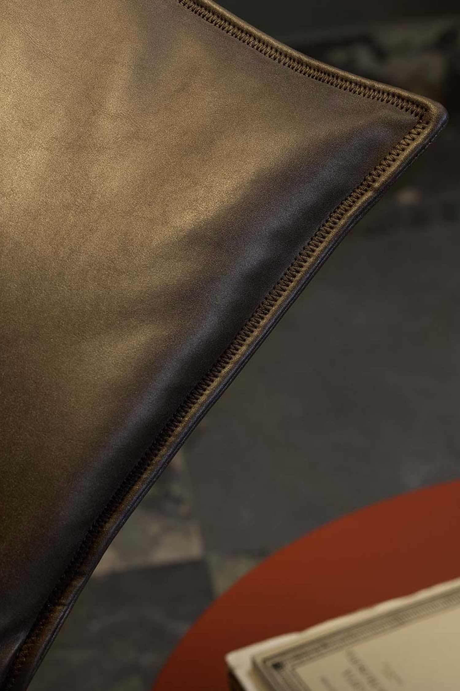A close-up, high-angle shot of the corner of a dark brown leather cushion, possibly from an armchair. The focus is on the diagonal stitching along the edge of the cushion. The background is blurred, showing a dark gray and red surface, suggesting a table and a dark floor.