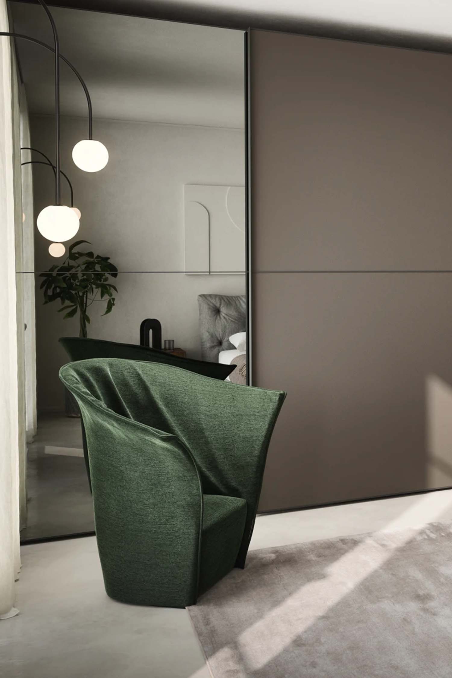 A large bedroom features a floor-to-ceiling wardrobe with a combination of dark gray panels and reflective mirror panels. One section of the wardrobe is open, revealing an organized closet interior with hanging clothes and shelves. In the foreground, a gray bed with a beige throw blanket is partially visible on the right. A unique dark green armchair with a high, rounded back is placed in the foreground to the left, and a large plant is visible in the reflection of the mirror. A modern black and gold lamp sits on a wooden nightstand on the far right.