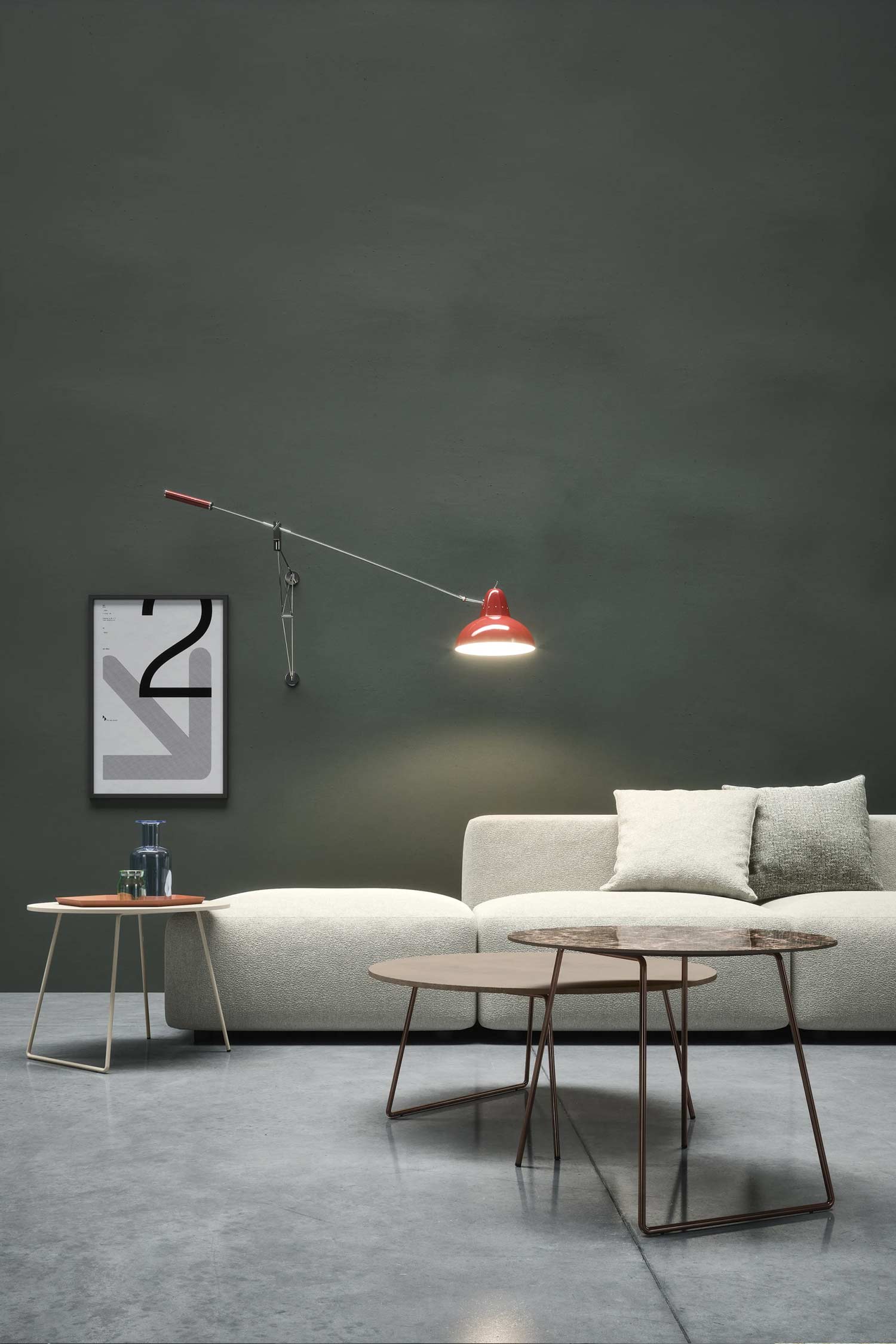 A low-angle shot of a minimalist living room with a cream-colored sofa against a dark green wall. In the foreground are two modern nesting coffee tables with marble tops, while a red-shaded wall lamp and framed art complete the scene.