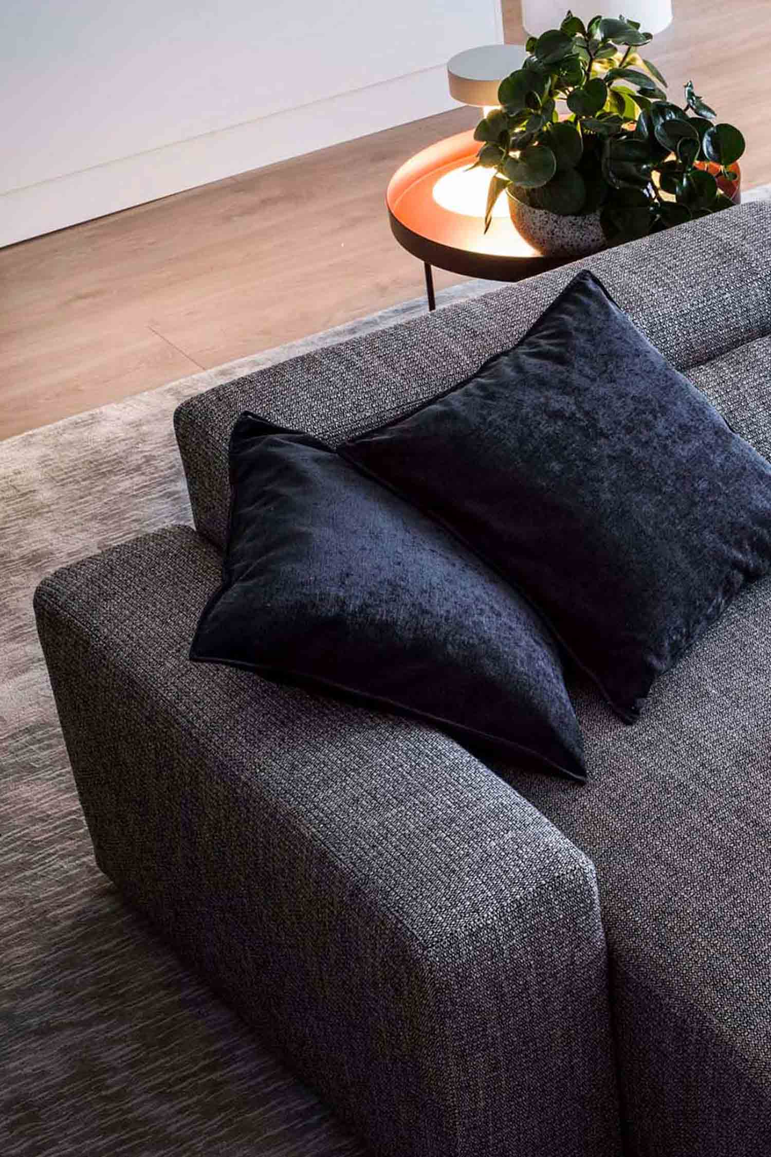 A close-up, high-angle shot of a gray fabric sofa. Two dark blue or black velvet pillows are on the armrest. A small side table with a plant and a lit lamp is in the blurred background.
