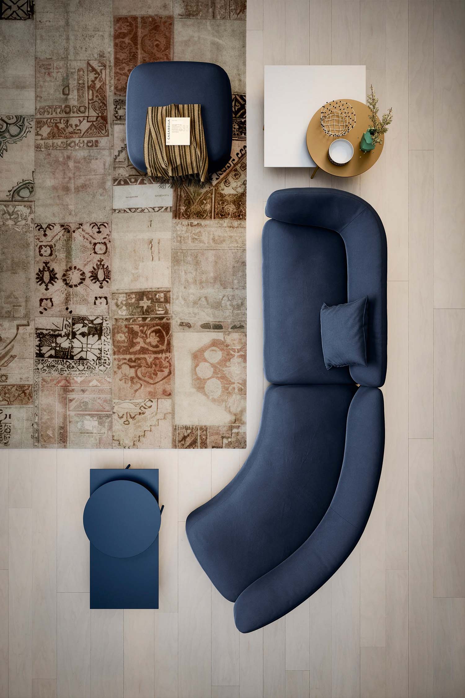 An aerial shot shows a modern living room setup. A blue, organically shaped sectional sofa with a single throw pillow is placed on a light wood floor. Next to it, a small, two-tiered circular blue table stands alone. A small, square blue ottoman with a book and a striped throw blanket rests on a large, distressed patchwork rug with patterns in earthy tones. A two-tiered, asymmetrical white and wood coffee table sits next to the sofa, holding a small decorative bowl, a vase with greenery, and a round woven placemat with a candle.