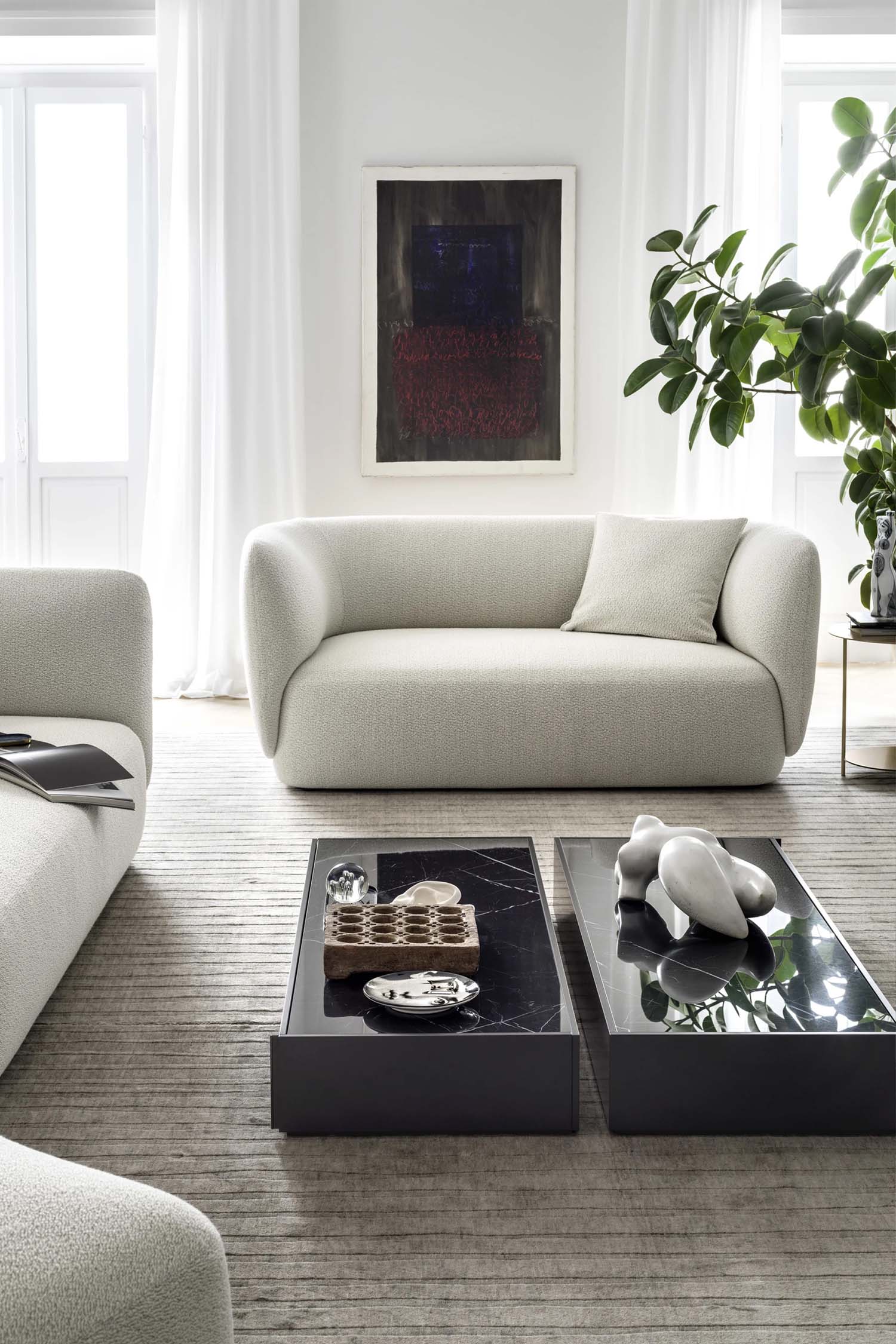 A close-up of a modern, curved sofa in a light bouclé-style fabric. The photo is taken from a side angle, showing the rounded edges of the armrest and backrest. A black metal bookshelf with a plant on top is next to the sofa, and a modern chandelier hangs in the background.