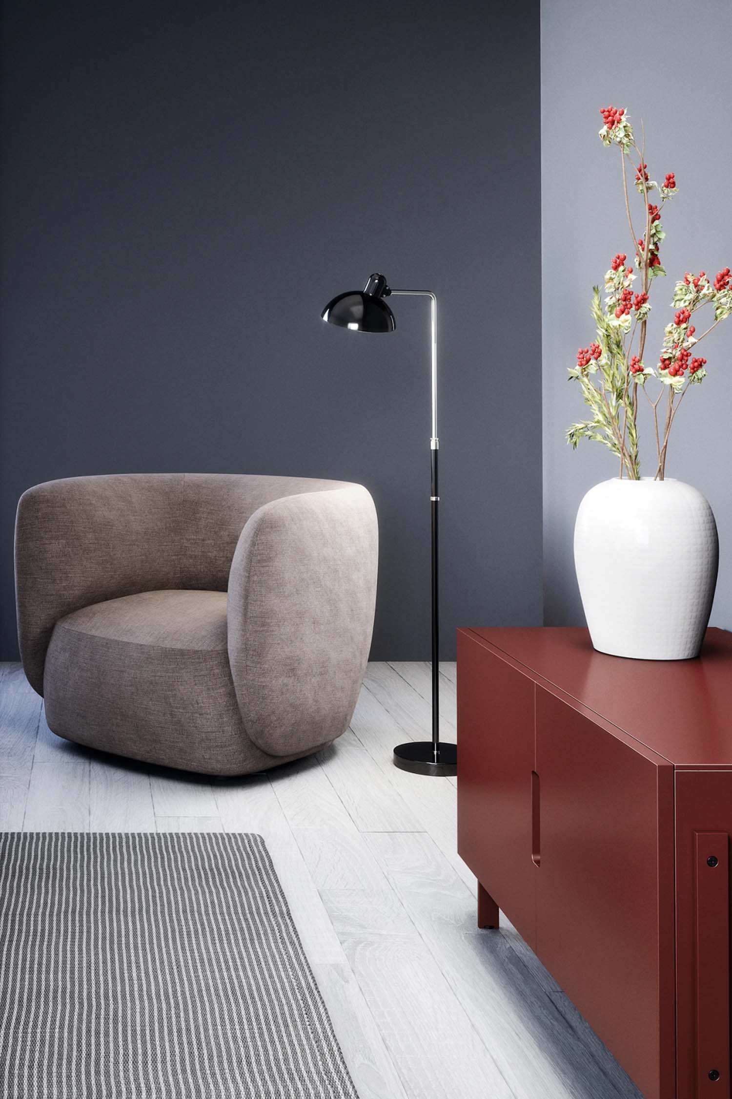 A cozy living space with a dark gray and light gray wall. A light taupe, curved armchair sits on a gray and white striped rug. A tall black metal floor lamp is positioned behind it. To the right, a long, brick-red console cabinet with a matte finish stands on black legs, holding a white vase with red and green berries and a white decorative number '14' sculpture. The floor is light-colored wood.