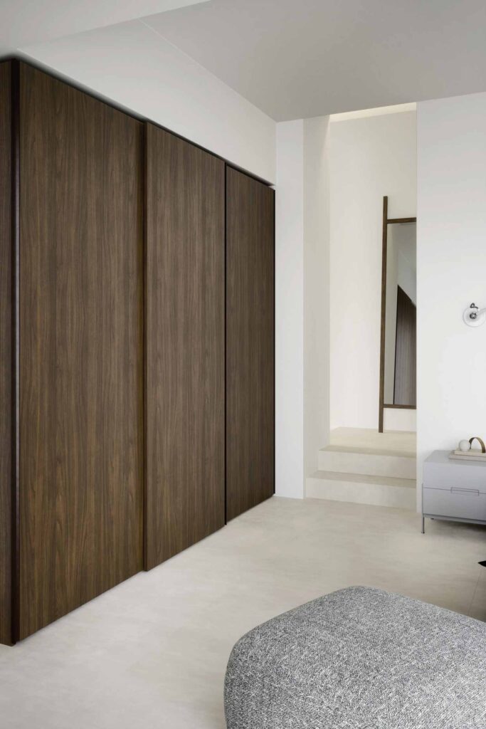 A wide-angle shot of a modern and minimalist bedroom interior. A large, built-in wardrobe with dark wood veneer sliding doors dominates the left side of the frame, spanning from floor to ceiling. In the foreground, a gray textured ottoman is partially visible. The room has light, almost white, flooring and clean white walls. In the background to the right, a narrow hallway or alcove leads to a full-length mirror leaning against the wall, reflecting the opposite side of the room. A small gray nightstand or dresser with a few decorative objects is against the wall on the right.