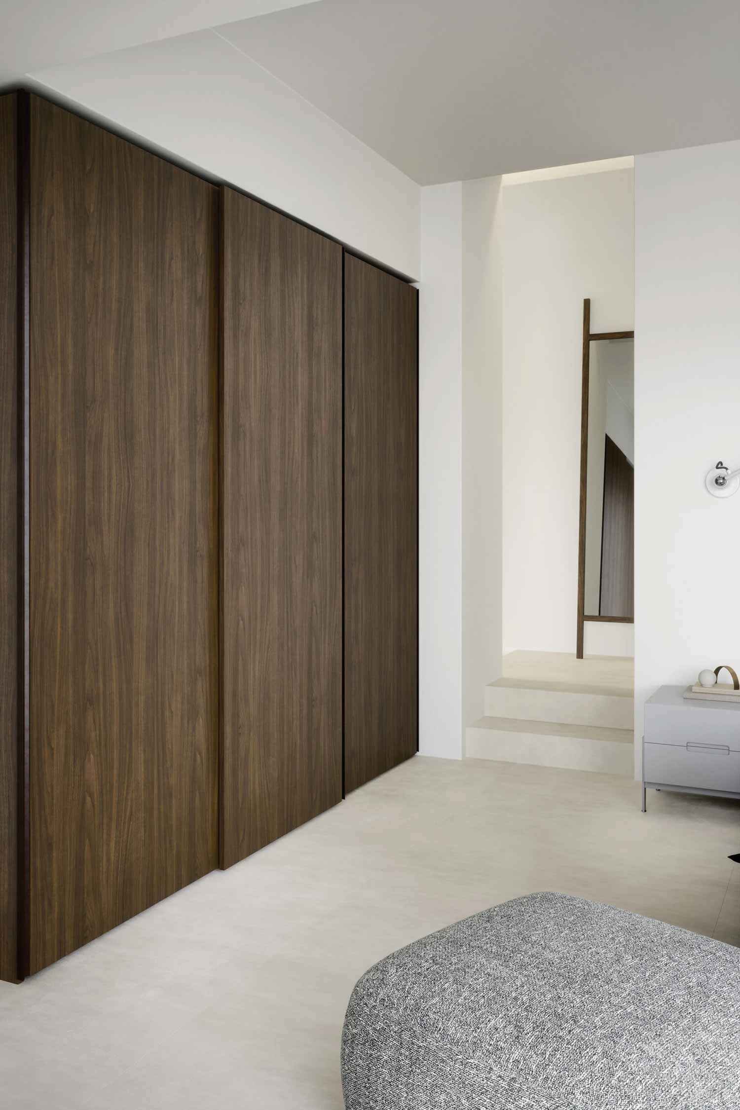 A wide-angle shot of a modern and minimalist bedroom interior. A large, built-in wardrobe with dark wood veneer sliding doors dominates the left side of the frame, spanning from floor to ceiling. In the foreground, a gray textured ottoman is partially visible. The room has light, almost white, flooring and clean white walls. In the background to the right, a narrow hallway or alcove leads to a full-length mirror leaning against the wall, reflecting the opposite side of the room. A small gray nightstand or dresser with a few decorative objects is against the wall on the right.