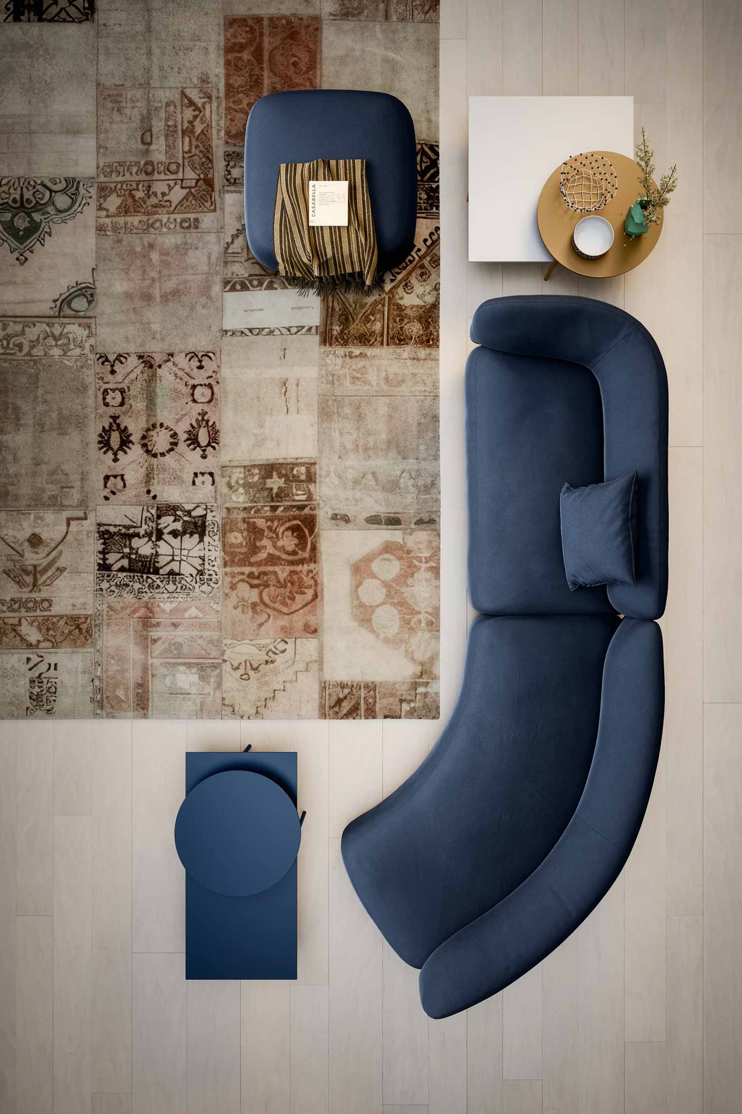 An overhead shot of a modern living room setup. A long, curvy, dark blue modular sofa with a small matching pillow is centered on the right side. A round, dark blue ottoman with a striped blanket and book is on the top left. A round side table with two tiers and a white top is next to the sofa, holding a bowl, vase, and wire basket. A small, square dark blue side table is on the bottom left. All furniture pieces are on or around a large, patchwork-style rug in various shades of brown and beige. The floor is light wood.