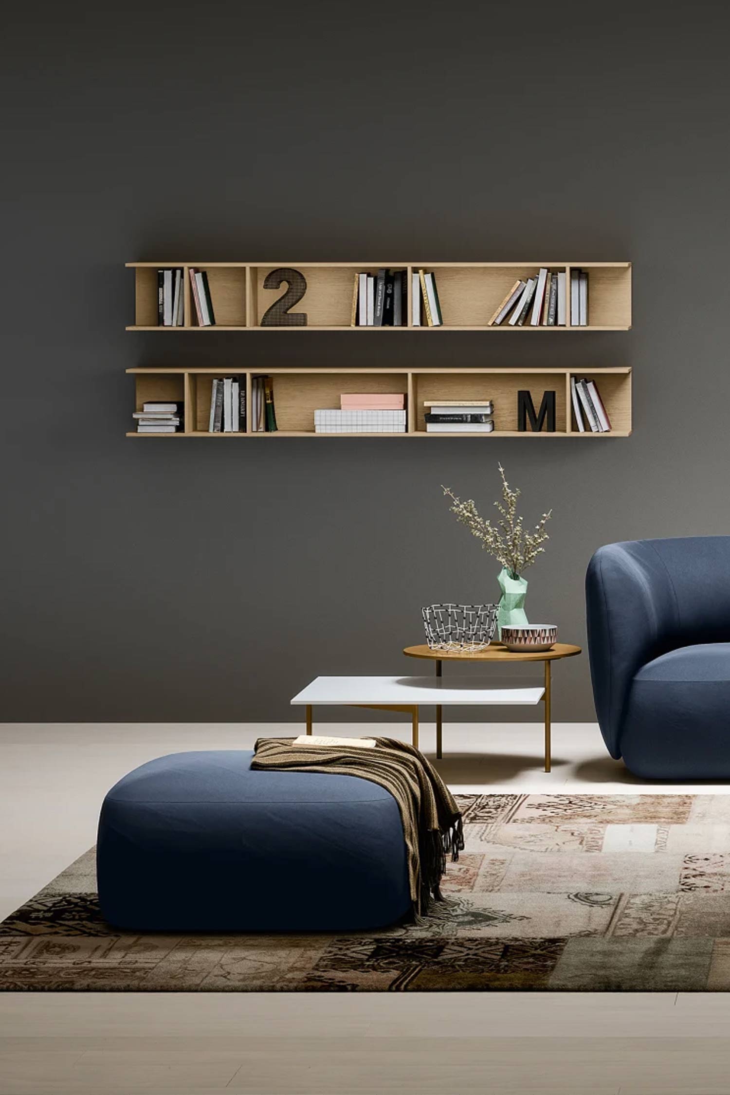 A full-height shot of a modern living room with a dark gray wall. A round, dark blue ottoman with a striped brown blanket on top is in the lower left corner, sitting on a patterned rug. Next to the ottoman, there are two small, tiered side tables. The top table has a white surface and the bottom one is a light bronze color. A light green vase with a few stems is on the bronze table, along with a wire basket. In the background, two long, light wood floating shelves are mounted on the wall, filled with books and a decorative number '2' and a letter 'M'. The right side of a curvy, dark blue sofa is partially visible. The floor is light wood.