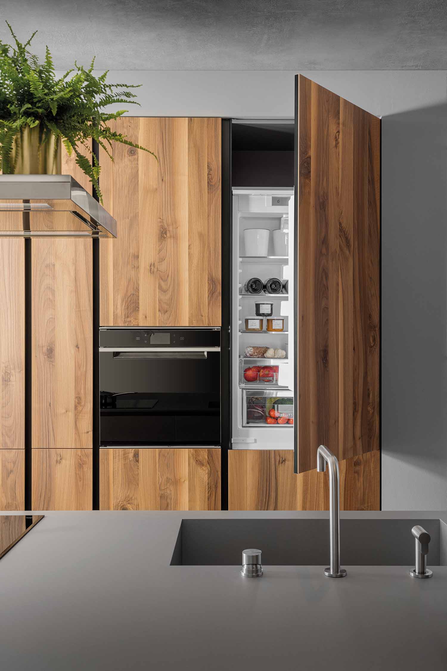 A close-up of a tall, wood-grain kitchen cabinet with its integrated refrigerator door ajar. The inside of the fridge is visible, with fresh food and bottles neatly organized. The surrounding cabinetry features a built-in oven, creating a sleek, unified look.