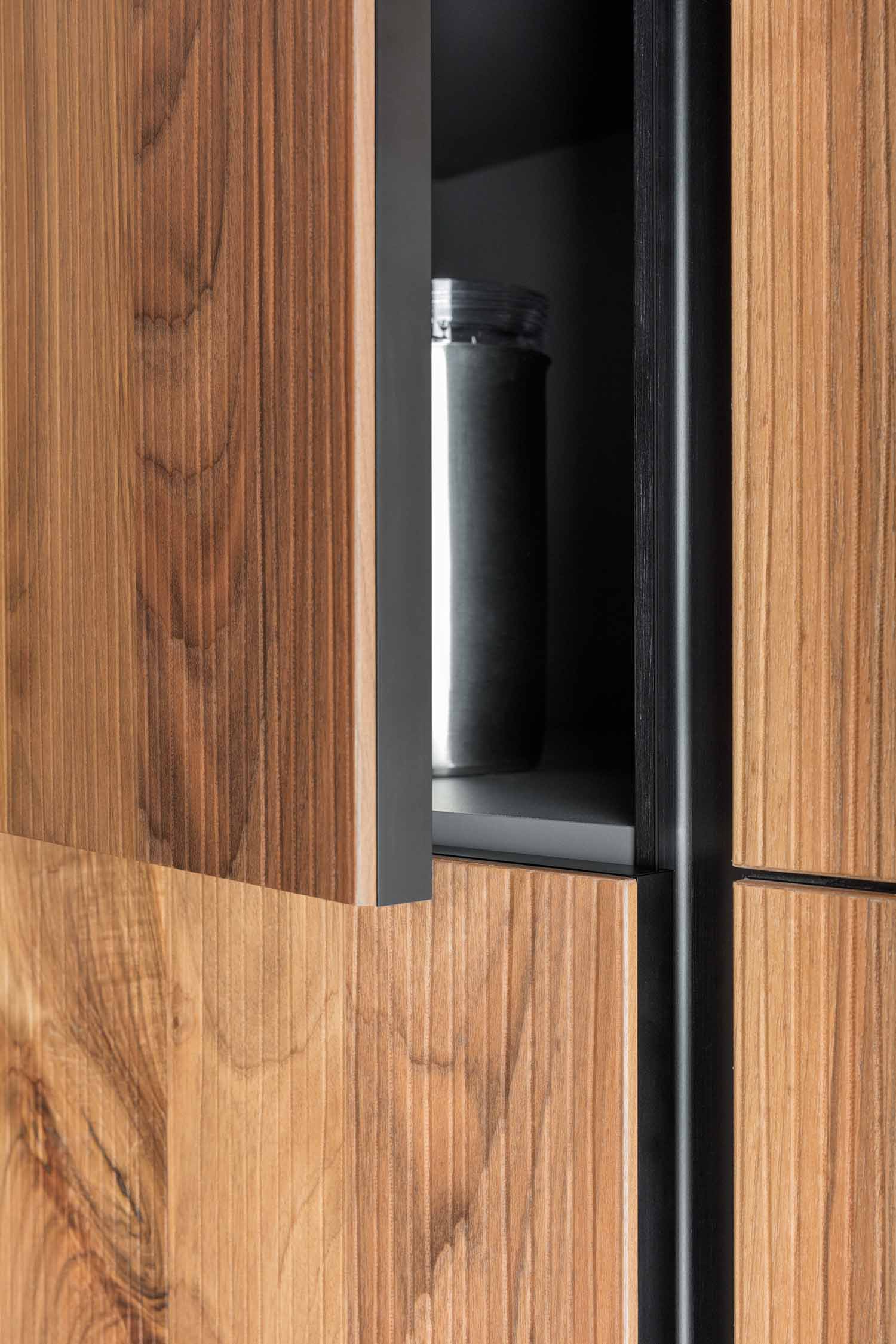 A detailed shot of the unique edge detail and handleless design of the kitchen cabinets. The image focuses on an open cabinet door, showcasing a dark, recessed vertical handle groove that allows for a seamless, clean look.