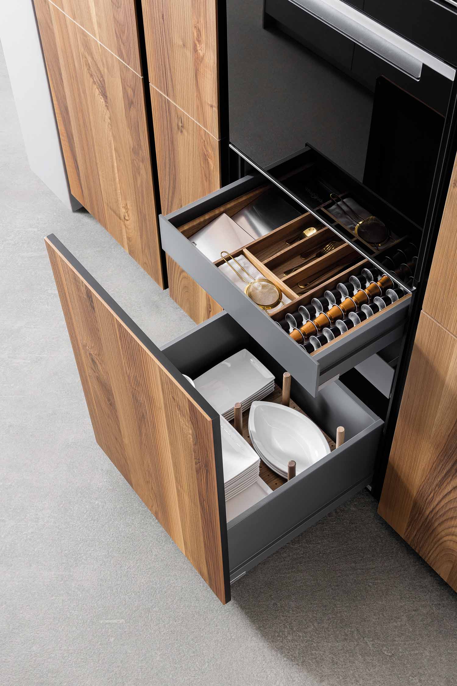 An image of two large, deep drawers in a wood-grain cabinet, opened to reveal their contents. The top drawer contains various utensils and cutlery in a wooden organizer, while the bottom drawer holds a stack of white plates and serving dishes, demonstrating smart storage solutions.