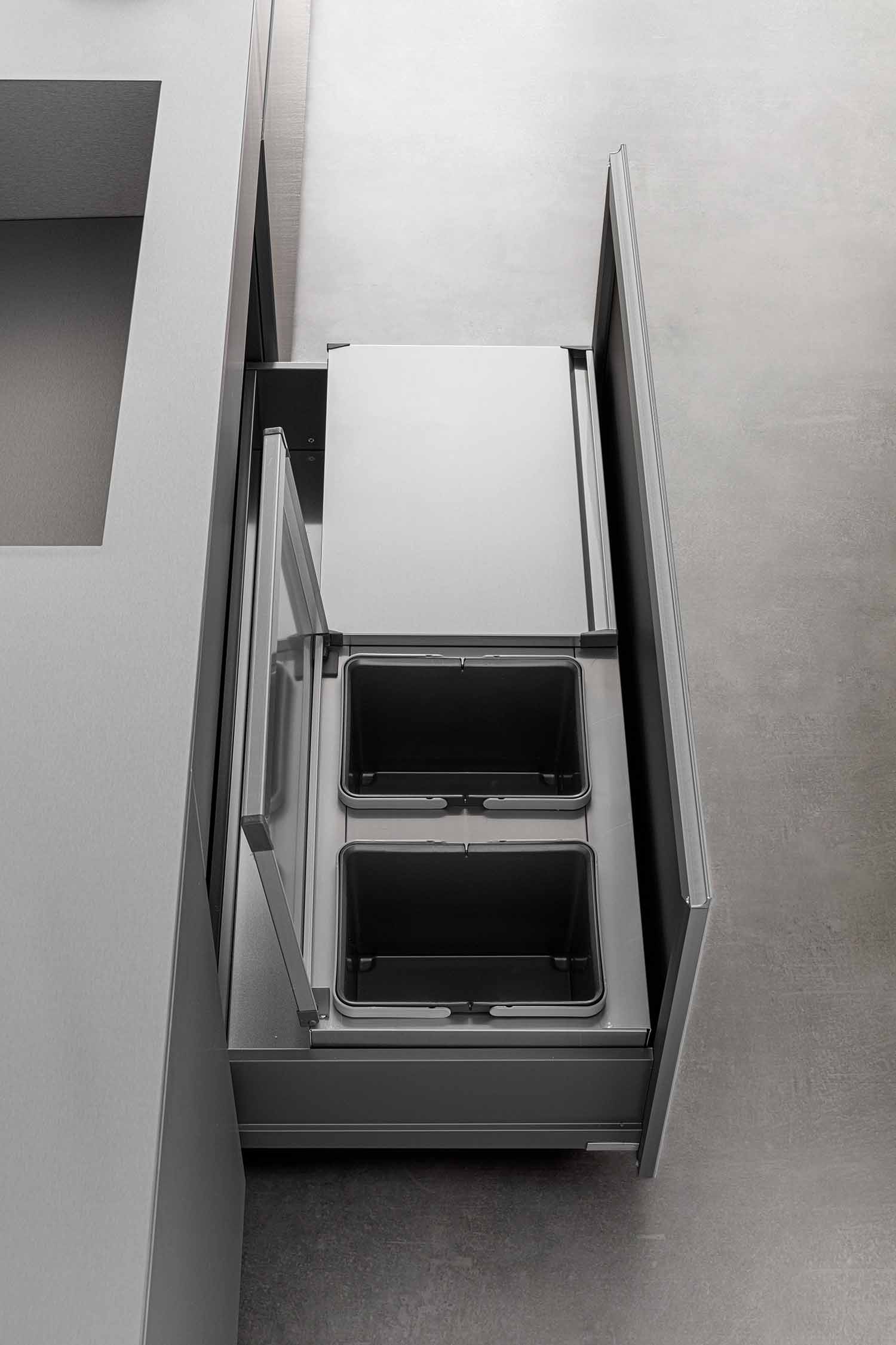 An overhead shot of a deep kitchen drawer with an integrated waste and recycling system. The drawer is pulled out to show two removable black bins for sorting waste, along with a flat metal lid to contain odors. The internal structure of the drawer is a clean, metallic grey.