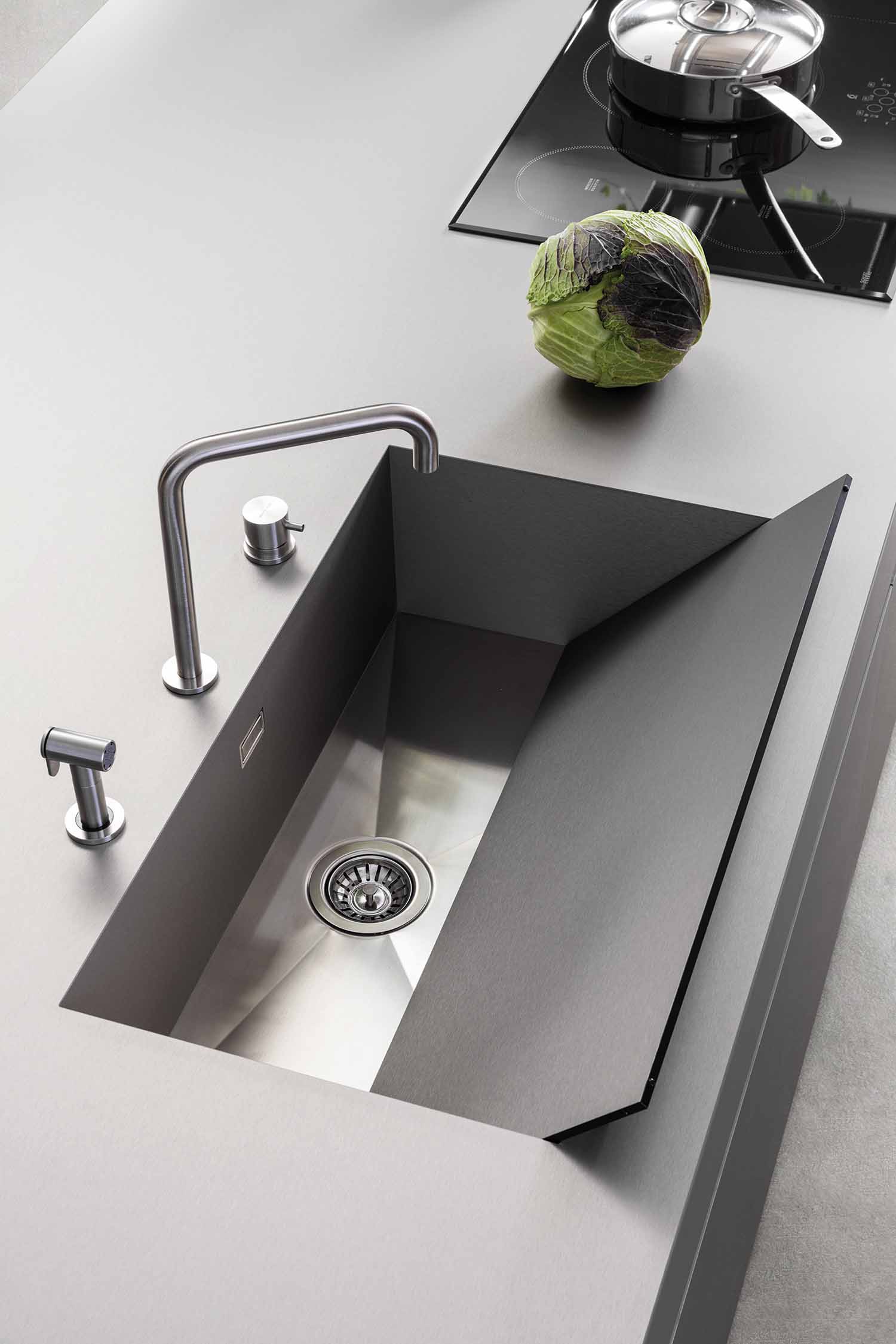 A close-up of the integrated kitchen sink on a grey, minimalist countertop. The sink is a seamless, modern design with an elegant stainless steel faucet and a matching secondary tap. A fresh cabbage is sitting on the counter near an induction hob.