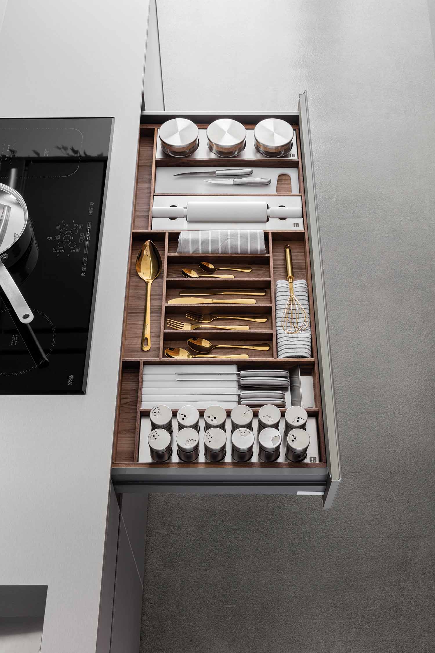 An overhead view of a perfectly organized kitchen drawer. The drawer, made of dark wood, is partitioned into sections holding various items, including a golden cutlery set, a white rolling pin, a whisk, and multiple spice jars.