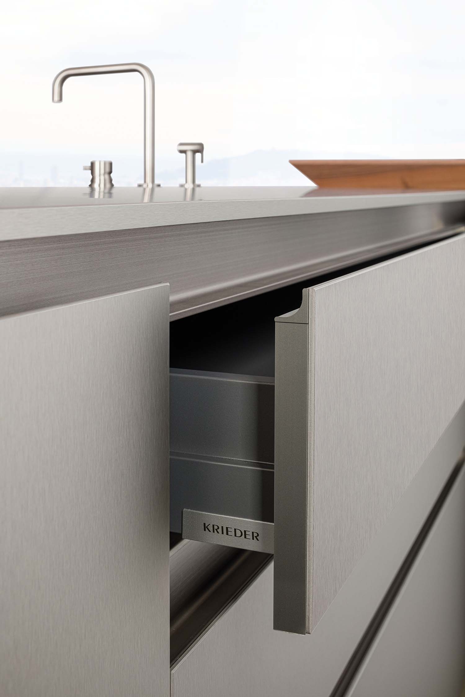 A side shot of a closed and an open drawer on the kitchen island. The open drawer reveals a minimalist metal handle pull with the brand name "Krieder" visible. The sleek, handles-free exterior and the fine brushed metal finish are emphasized.