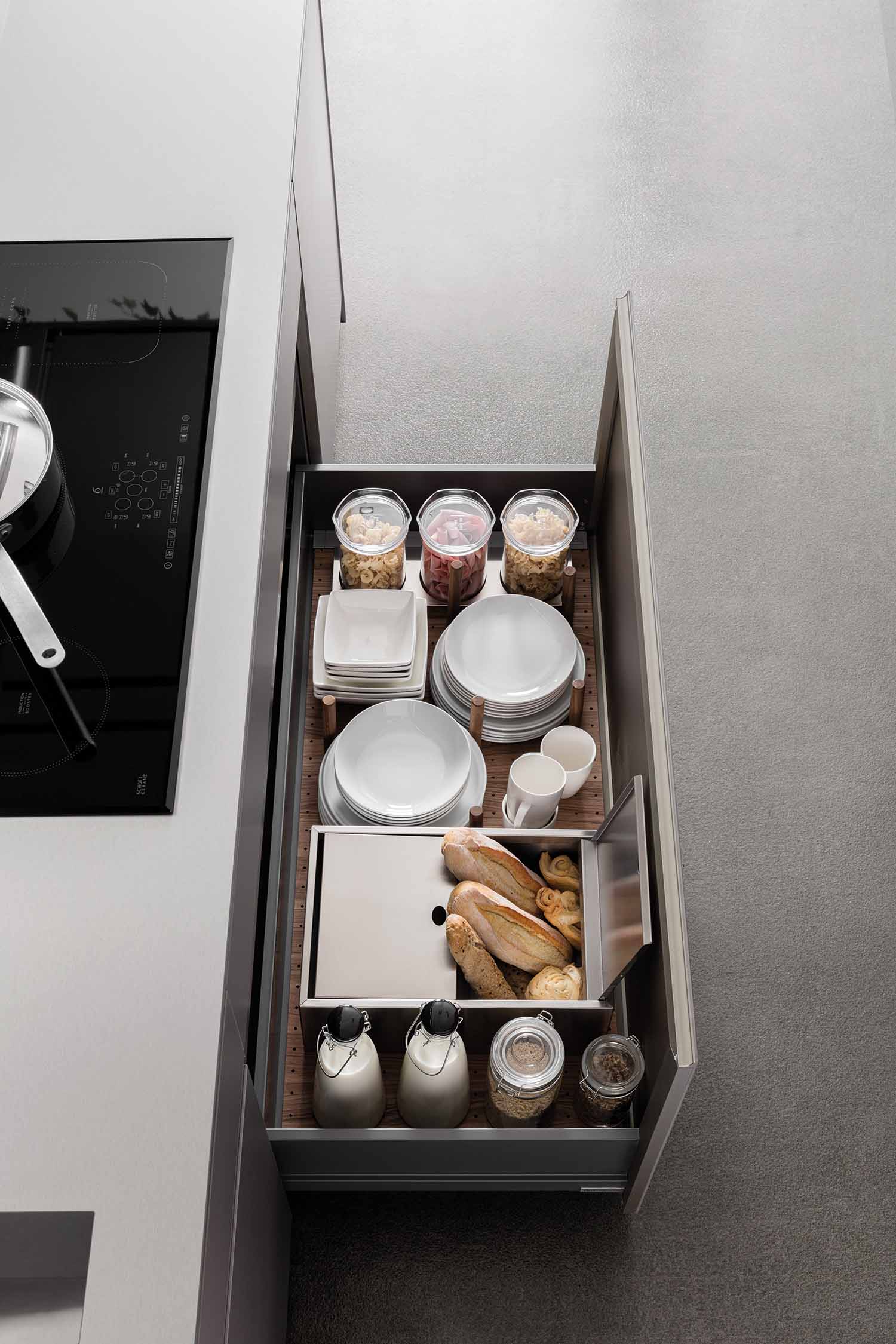A top-down view of a large, open kitchen drawer with an organized storage system. The drawer holds stacks of white plates, bowls, and mugs, along with several glass jars of dry goods and a separate compartment for bread. The interior has a dark wood-like base with lighter dividers.