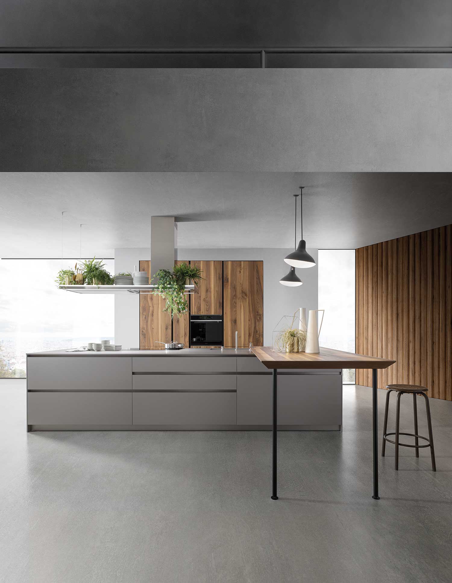 An overview of a modern open-plan kitchen and dining area. A long, grey, minimalist kitchen island dominates the space. A raised extractor hood with built-in planters hangs above the island. A wooden table with a single leg extends from the island, and a black bar stool is tucked underneath. The background features a striking wood-slatted wall and a built-in oven unit.