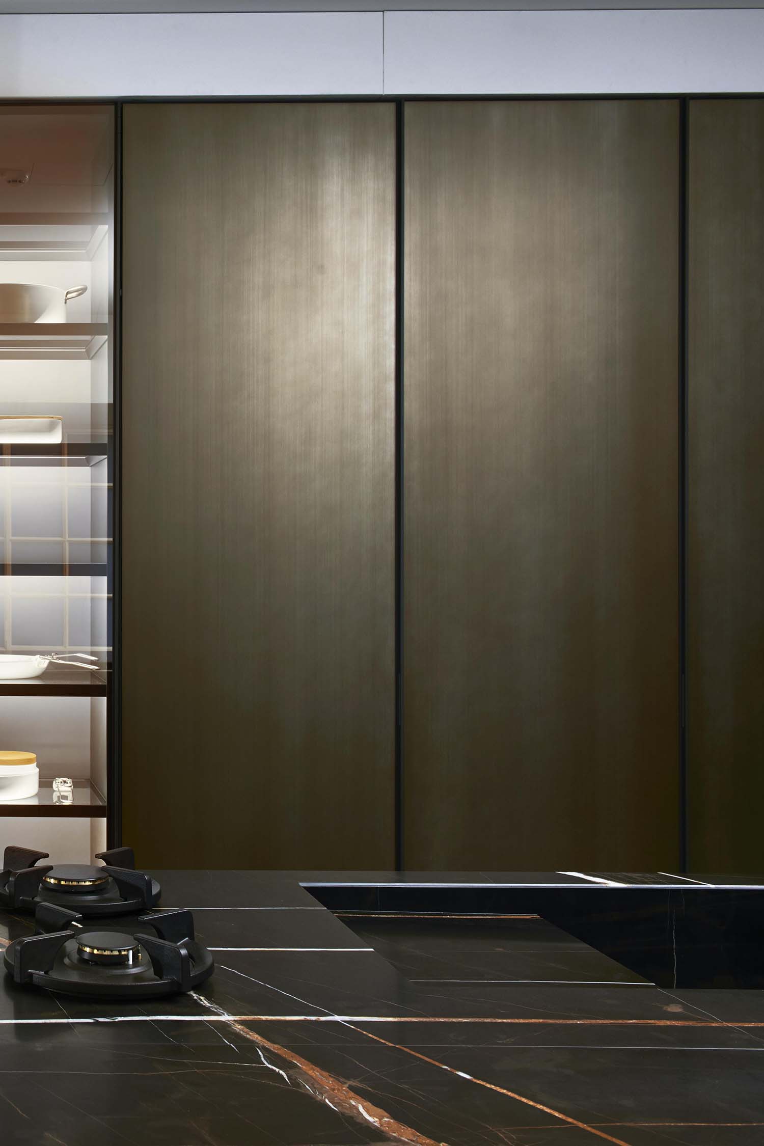 A close-up, low-angle view of an open kitchen cabinet revealing a bespoke wooden bottle storage solution. The cabinet has an integrated light strip at the top, illuminating the bottles within.