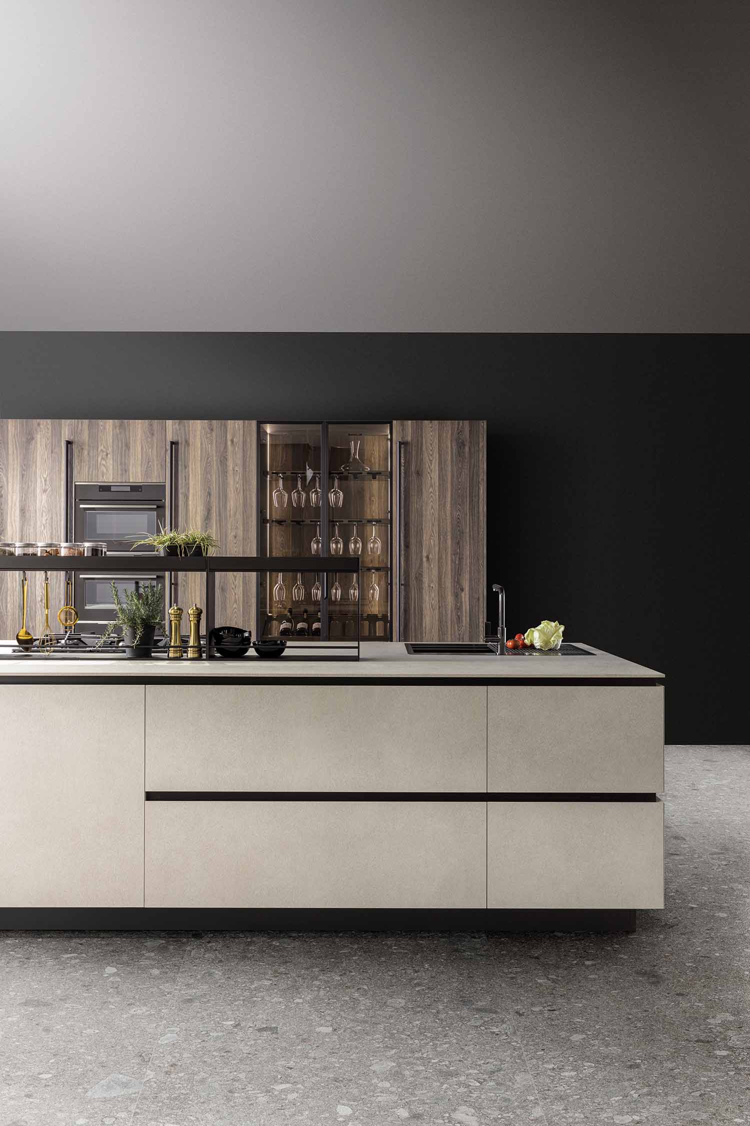 A view of a modern kitchen island with a beige, textured finish. A gas hob is integrated into the countertop, along with a sink and a black faucet. A raised black metal frame sits on the island, holding various kitchen tools and plants. The backdrop is a black wall and a set of tall, wood-grain cabinets with a built-in oven and a glass wine display.