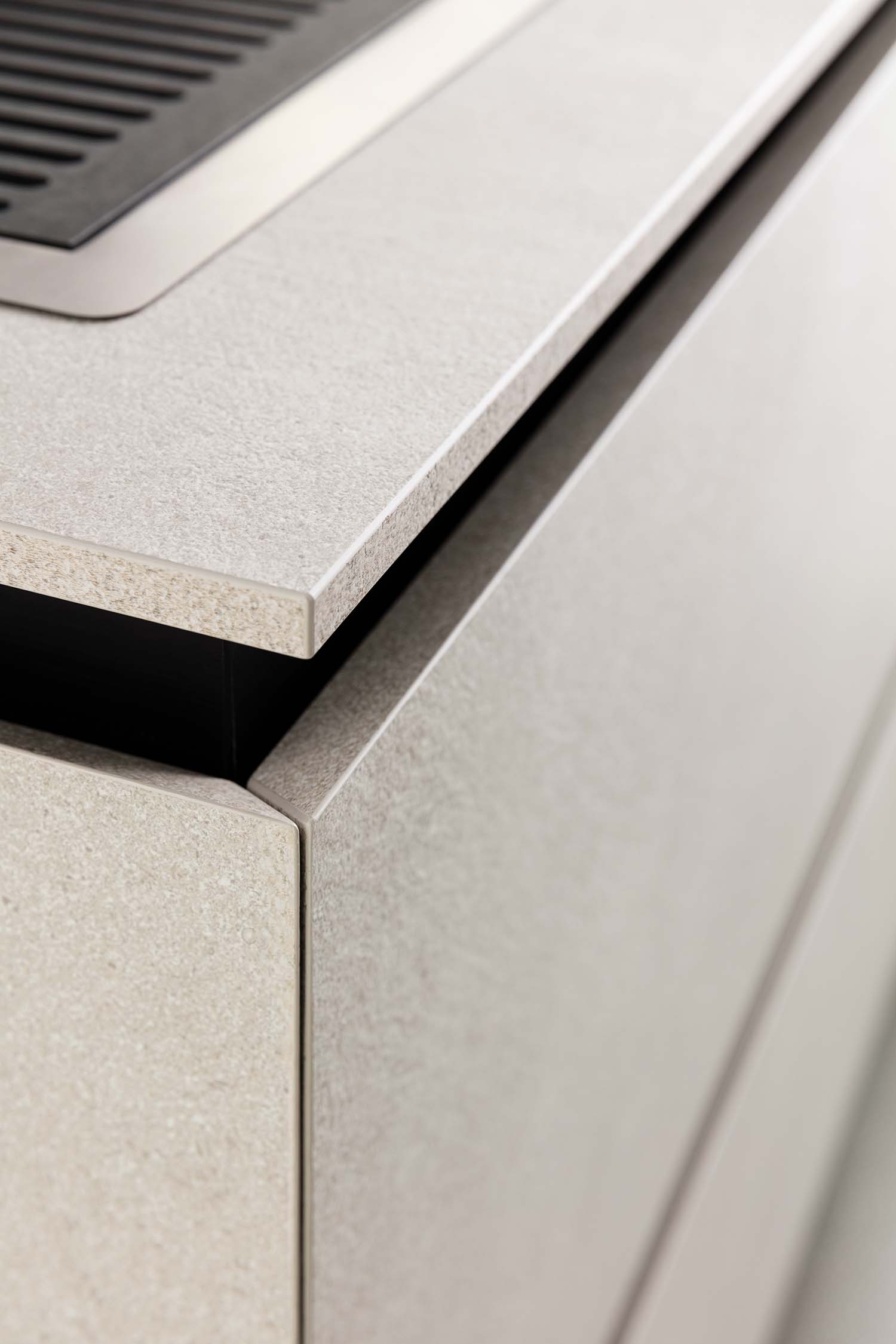 A close-up of the handleless design of the kitchen cabinets. The shot highlights the black "groove" or channel that serves as a pull for the drawer, providing a clean, minimalist look. The countertop is a textured, light beige material.
