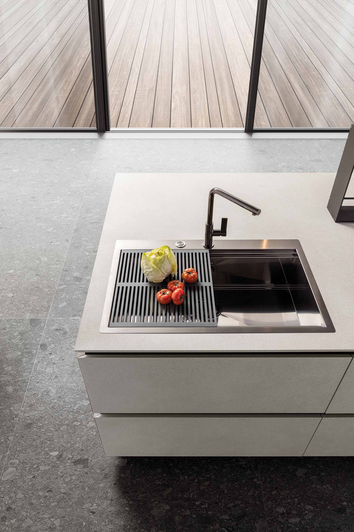 A top-down view of the kitchen island's integrated sink area. The sink is made of stainless steel, and a black grooved rack sits over a portion of it, providing a versatile work surface. A shiny black faucet and a few fresh vegetables are on the countertop.