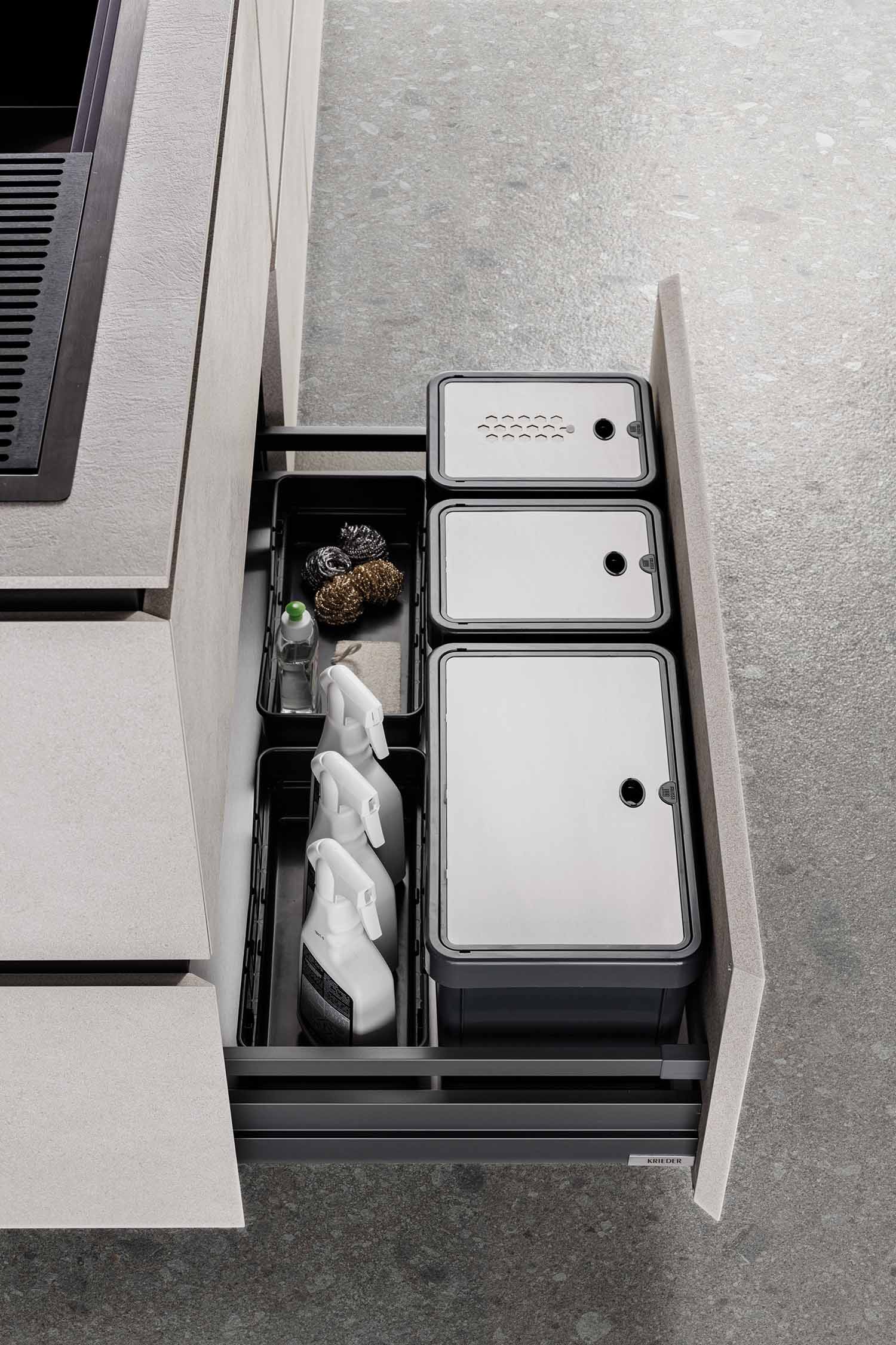 A view of a kitchen drawer opened to reveal an organized waste and recycling system. The black plastic bins are designed to fit neatly within the drawer, with different compartments for cleaning supplies and waste.