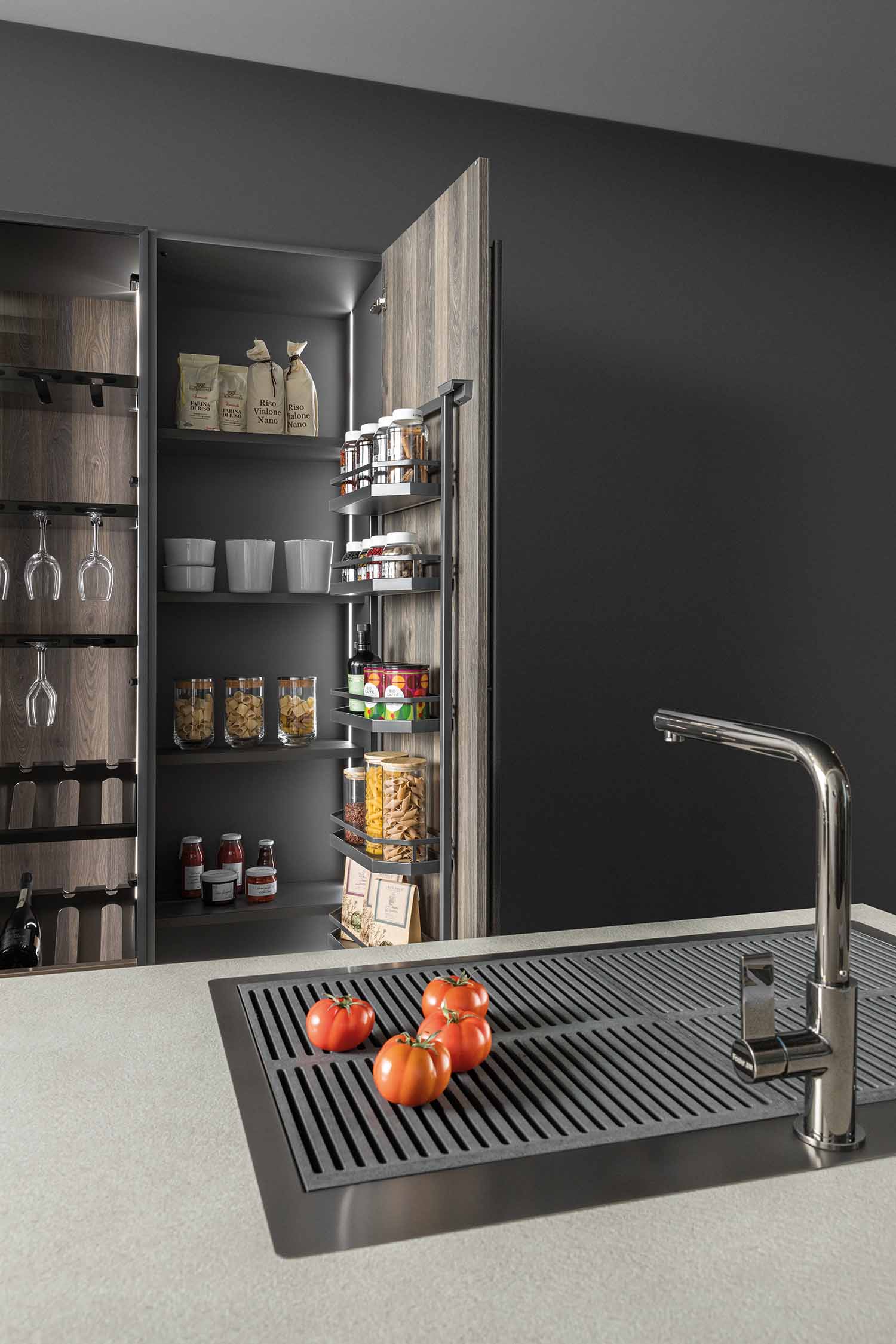 An image showing a tall pantry cabinet with a pull-out spice rack and adjustable shelves. The cabinet, made from a wood grain laminate, is lit from within, showcasing a variety of food items and glassware. A part of the kitchen sink is visible on the right.