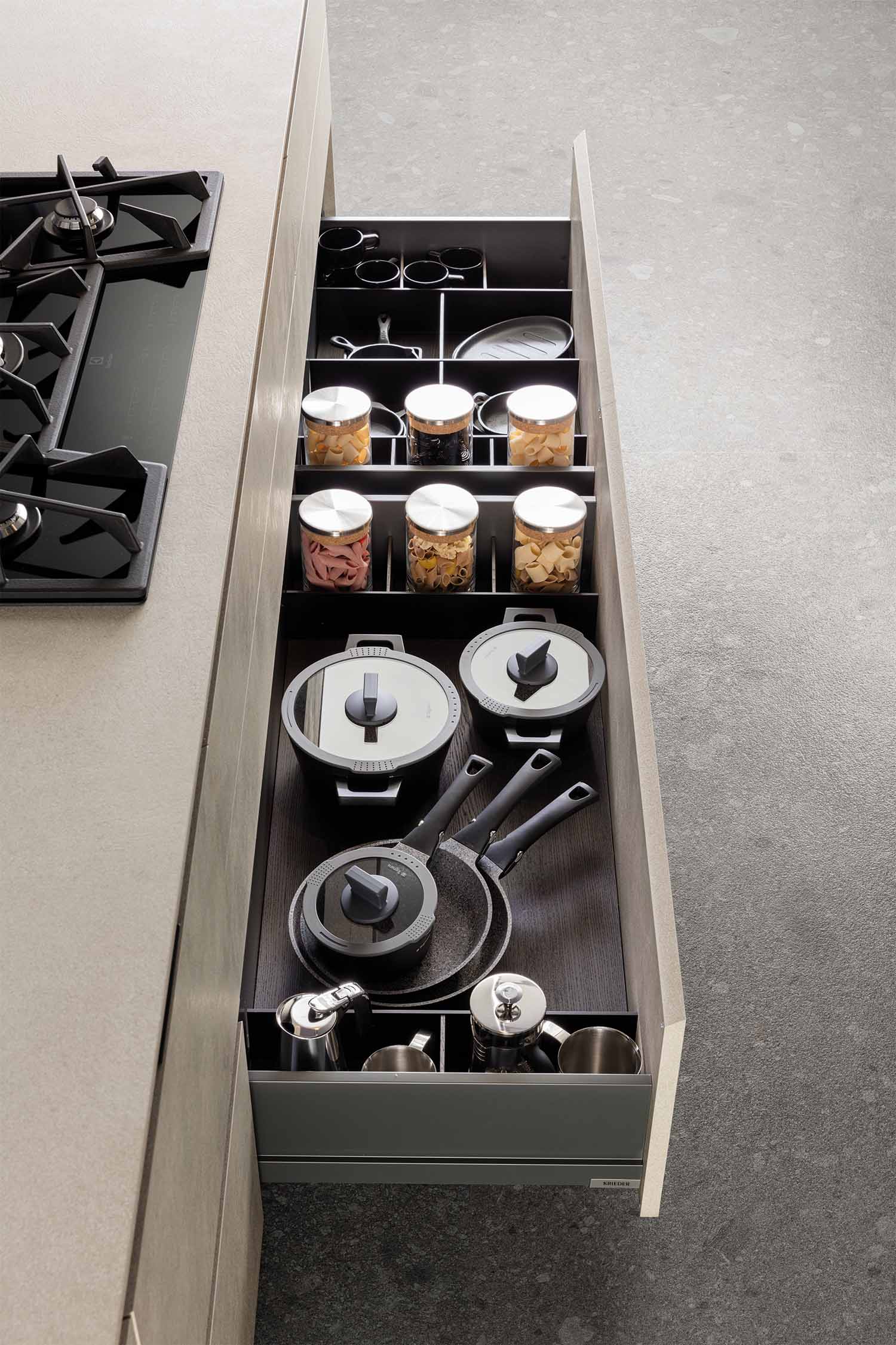 A top-down view of a large, deep kitchen drawer. The drawer is organized with modular dividers, holding a collection of pots, pans, and glass jars. The dark interior contrasts with the light, textured finish of the drawer face.