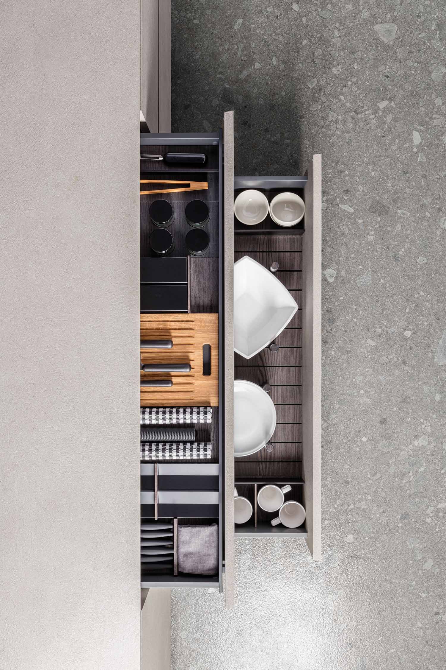 An overhead shot of two open, adjacent kitchen drawers. The left drawer contains organized cutlery, kitchen tools, and a wooden cutting board. The right drawer holds various sizes of white plates and bowls. Both drawers feature dark, modern dividers for optimal storage.