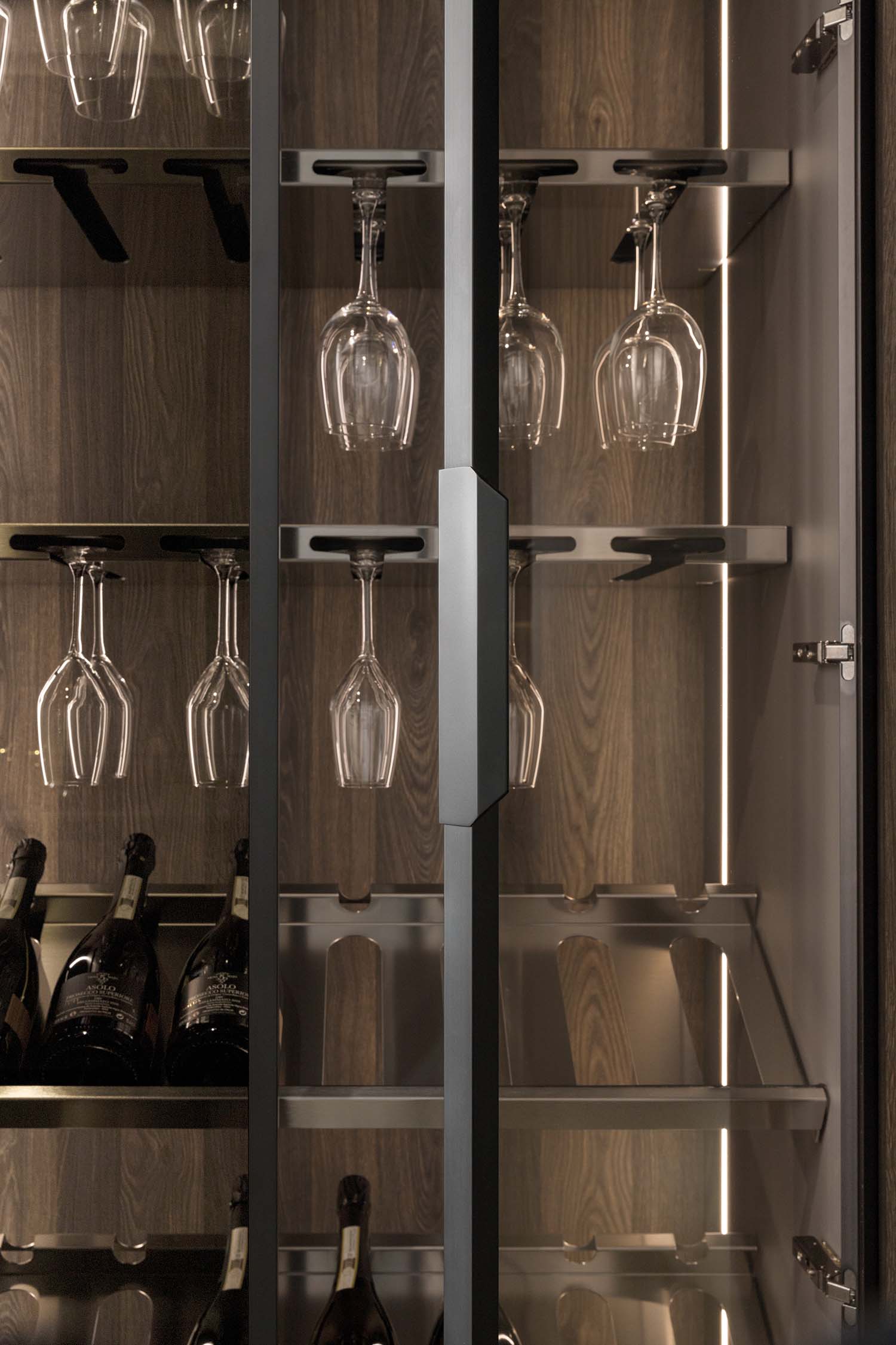 A close-up of the interior of a tall, glass-fronted wine cabinet. The cabinet has built-in stainless steel racks for wine glasses and shelves for bottles. LED lighting strips run vertically, illuminating the contents.