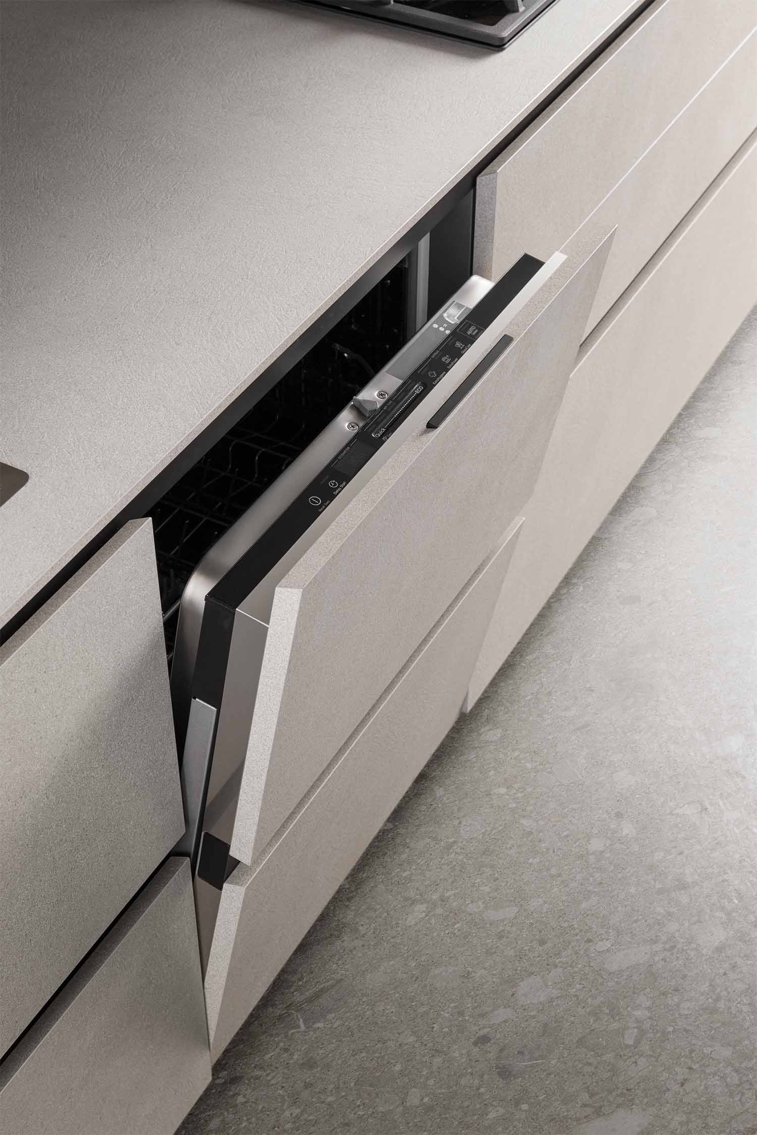 A detailed shot of a partially opened dishwasher, seamlessly integrated into the kitchen cabinetry. The front panel of the dishwasher matches the light, textured finish of the cabinets, maintaining the handleless aesthetic. The control panel is visible inside the door.
