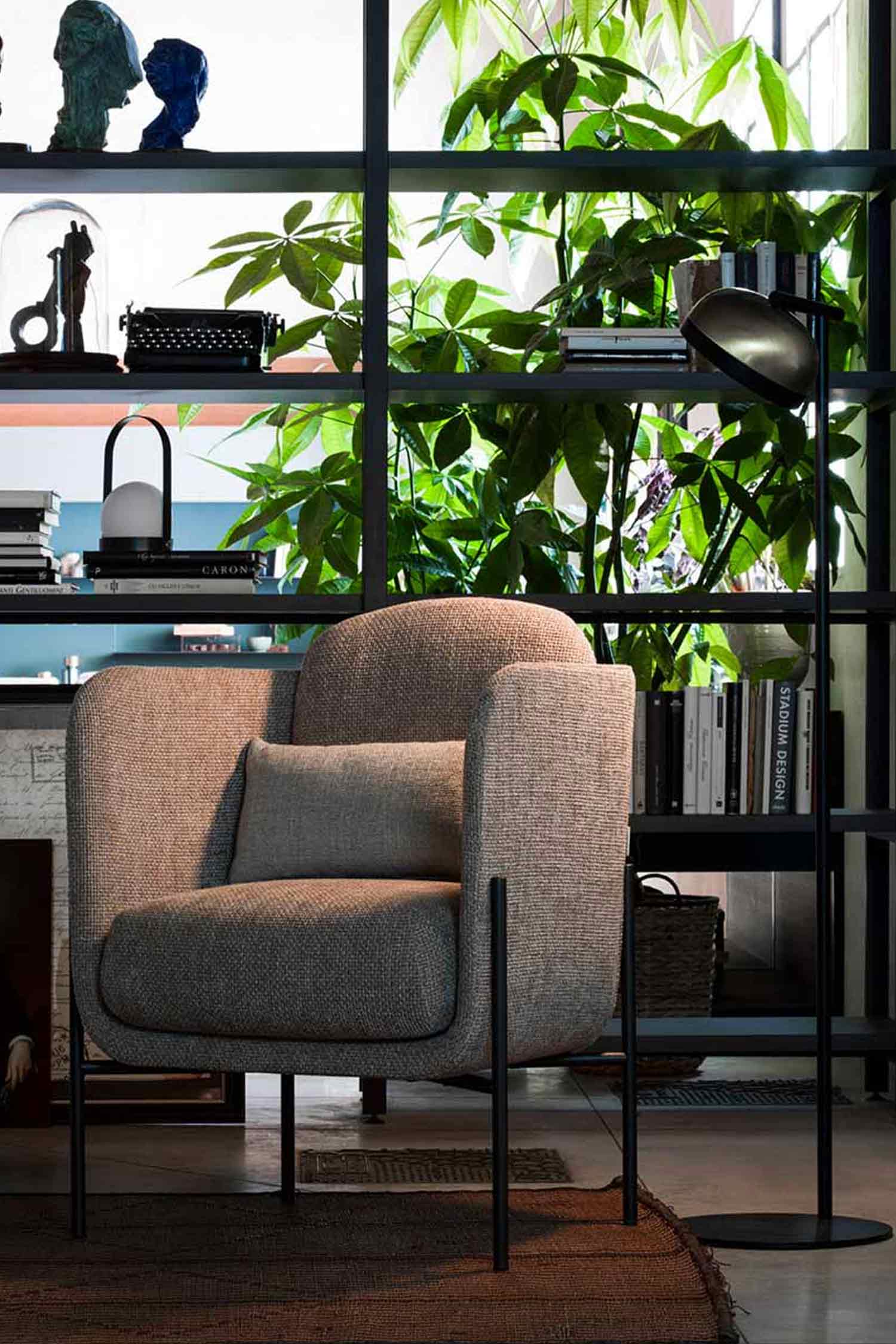 A professional, close-up shot of a modern armchair. The armchair has a light gray textured fabric, a plush back and seat, and a small, rectangular throw pillow. The chair sits on thin, black metal legs. Behind the armchair, a large, black metal shelving unit is filled with books, decorative sculptures, and lush green plants. To the right of the armchair, a modern floor lamp with a metallic, half-moon-shaped shade is positioned. The room has dark wood flooring and a small rug under the chair.