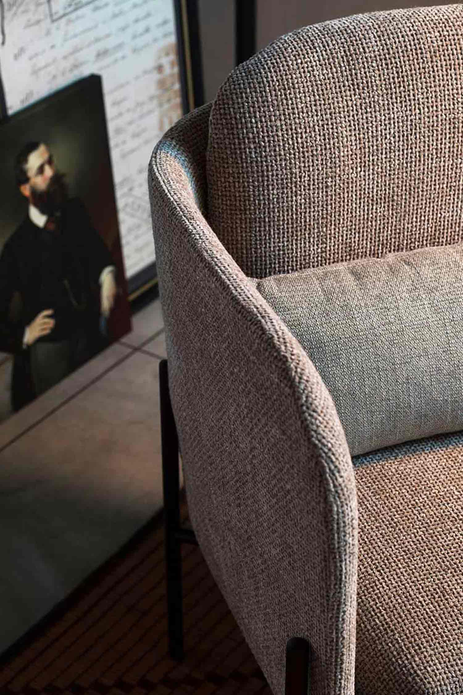 A close-up, high-angle shot of a modern armchair with a textured, light brown or beige fabric. The curved armrest and backrest of the chair are in sharp focus, showing the detail of the woven material. A cylindrical pillow in a lighter, contrasting fabric is propped against the back of the chair. In the blurred background, a framed portrait of a man with a beard, dressed in dark clothing, is visible. The floor is a dark wood, partially covered by a textured rug.