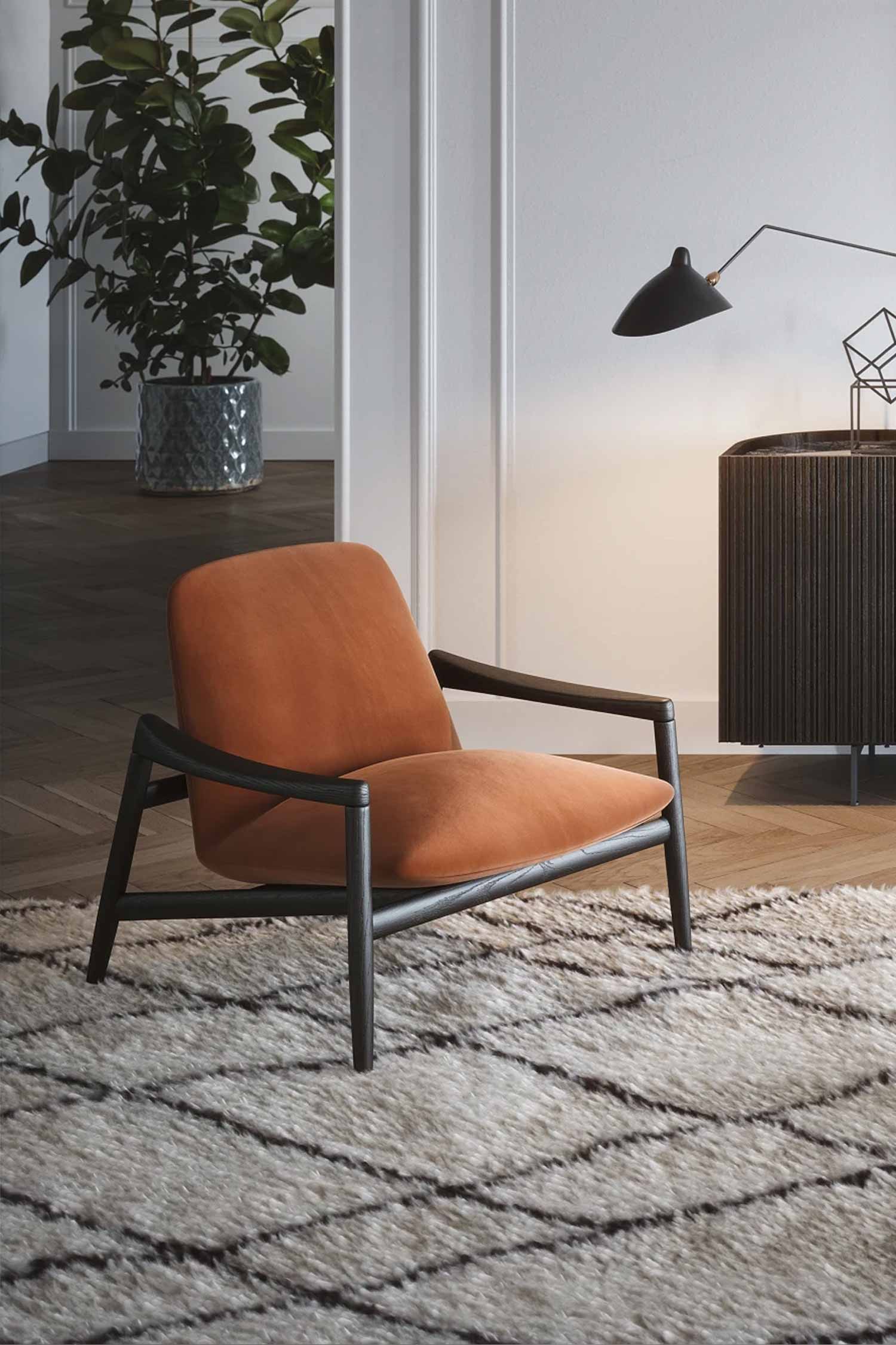 A modern and stylish living room features an orange upholstered armchair with a dark wood frame and armrests. The chair is positioned on a cream-colored, shaggy rug with a gray geometric pattern. In the background to the right, a dark wood credenza with vertical grooves is against a white wall. A black wall-mounted lamp illuminates the area above the credenza. A large potted plant is visible in a corner on the left. The room has light wood flooring.