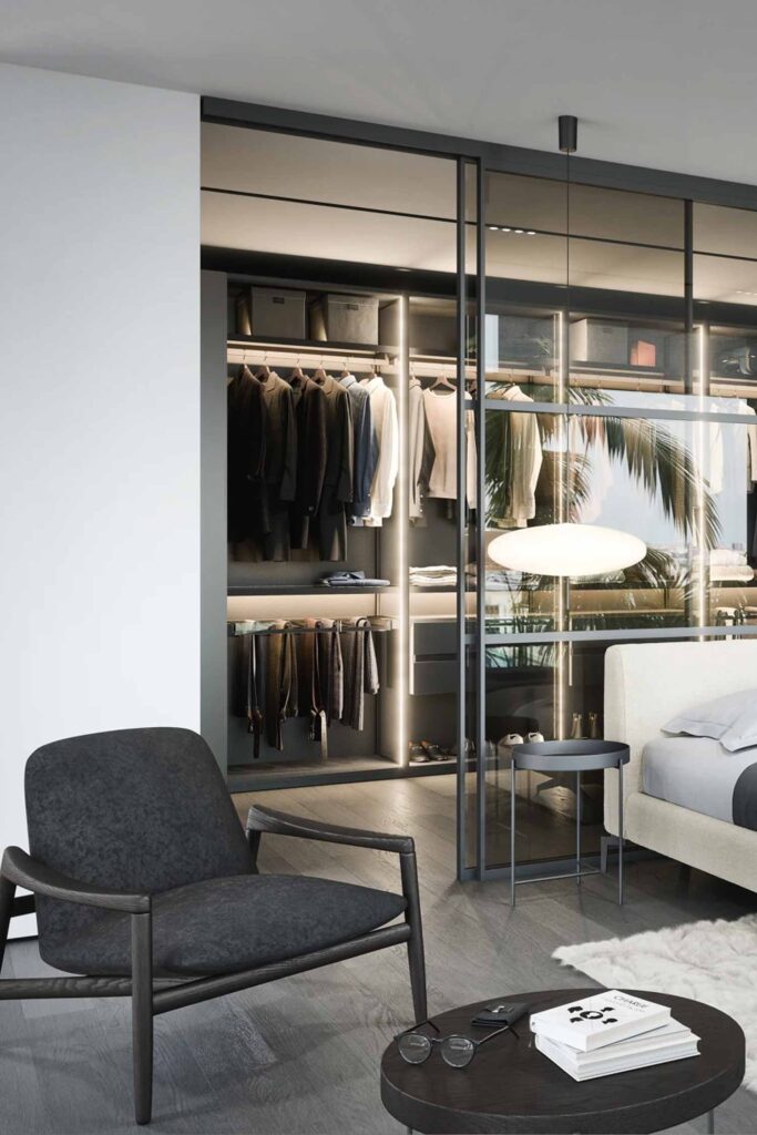 A low-angle, eye-level shot of a modern bedroom with a luxurious walk-in closet. In the foreground, a dark gray armchair with a wooden frame sits on light wood flooring. A round, dark wood side table with books and glasses is next to the armchair. Behind it, a large, glass-partitioned wall reveals an illuminated and organized closet, with clothes hanging on racks, folded items on shelves, and various storage solutions. The glass partition partially reflects an outdoor scene with palm trees. To the right, a bed with a light-colored upholstered frame and gray bedding is visible, with a small round side table next to it. A shaggy white rug is under the bed.
