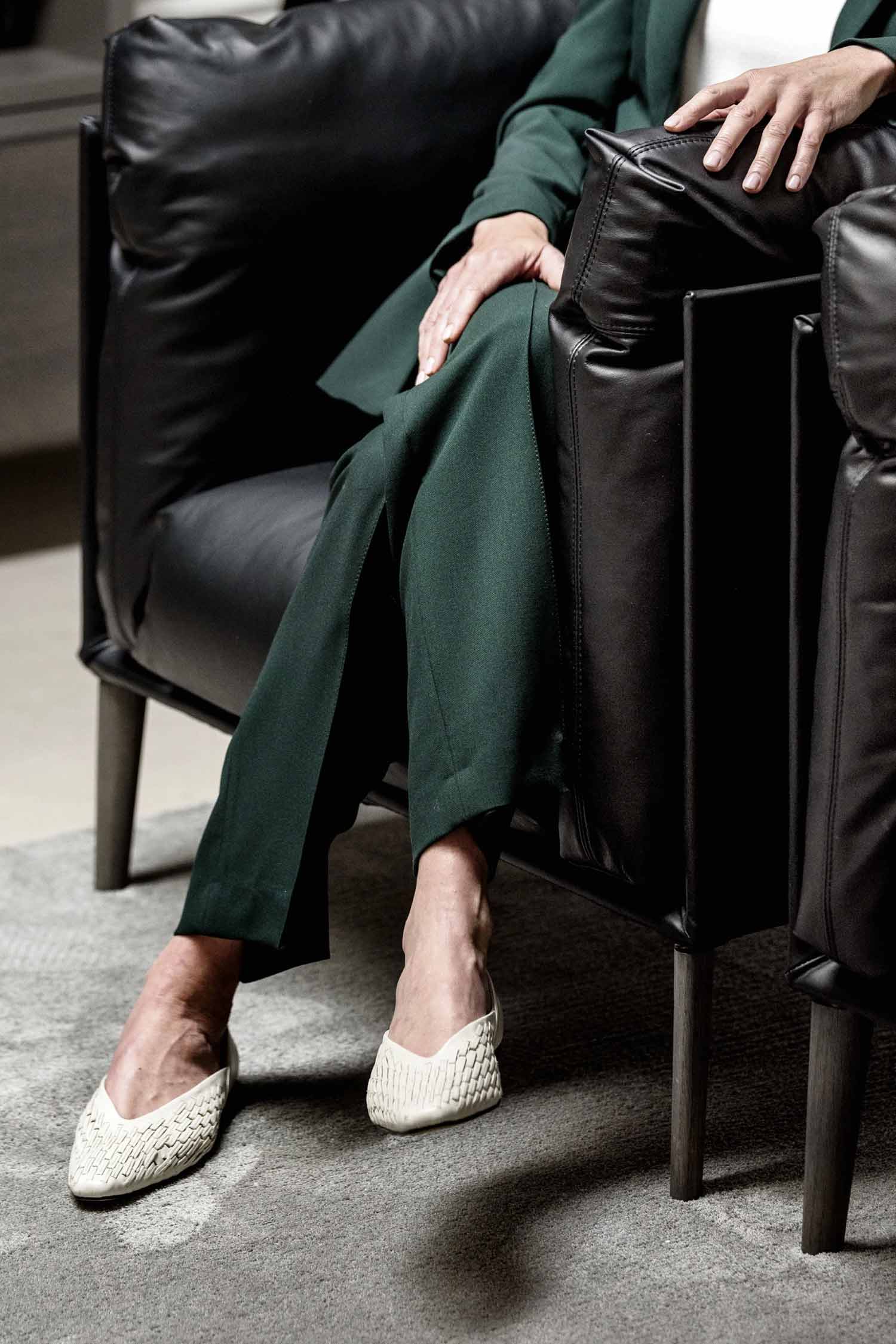 A person is seated in a black leather armchair, wearing dark green trousers and textured, off-white low-heeled shoes. Their hands are resting on their lap and the armrest. The armchair has black wooden legs, and a corner of a gray patterned rug is visible beneath it. The background is a blurred dark room.