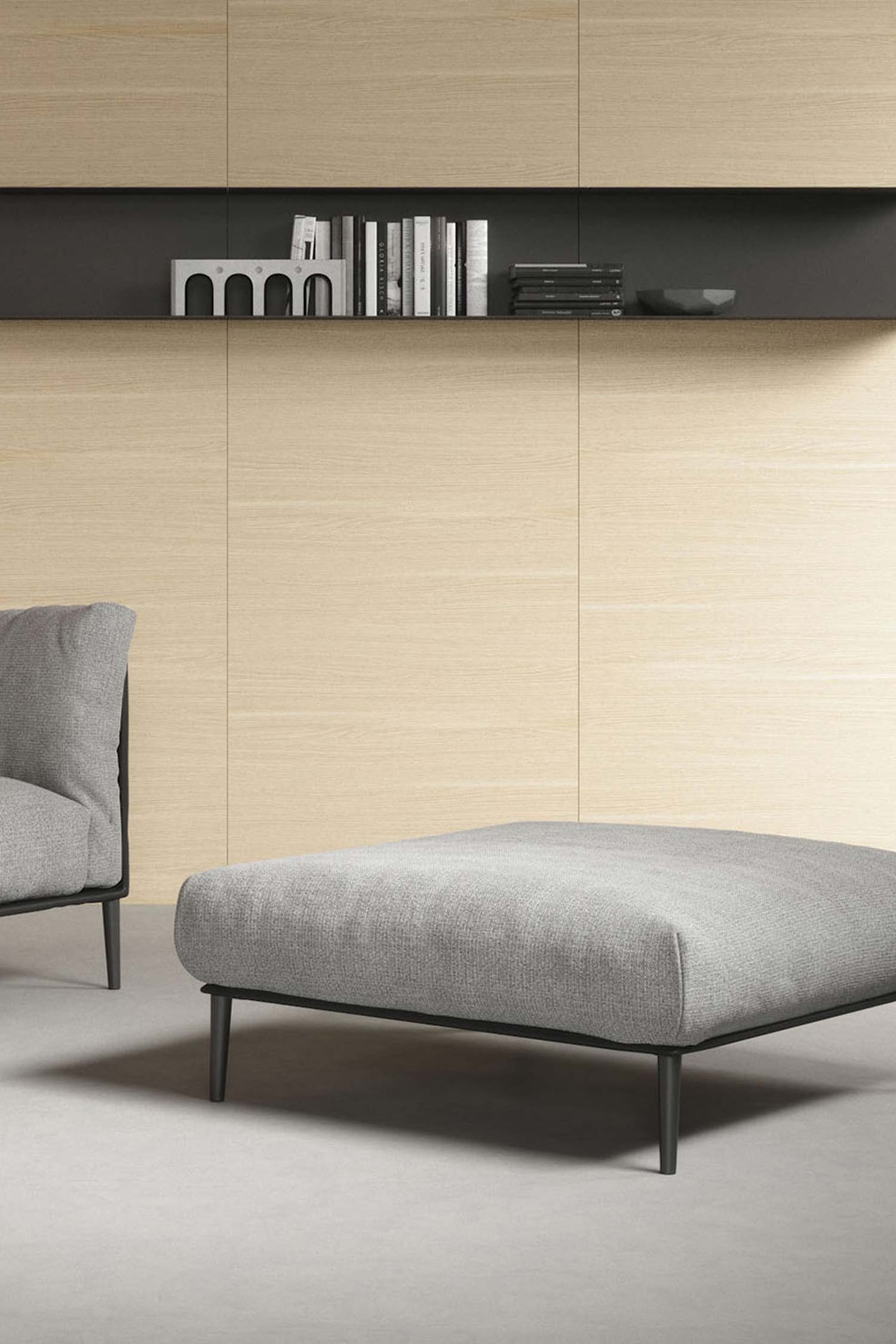 A low-angle shot of a modern living room or bedroom. A gray armchair with a black metal frame and legs is in the foreground, facing a light gray rectangular ottoman. Both are upholstered in a light gray fabric. In the background, a wall is covered in light wood-paneled tiles, with a long, black floating shelf holding books and decorative objects in the center. A black and white abstract painting is visible on the far left. The floor is light gray concrete.