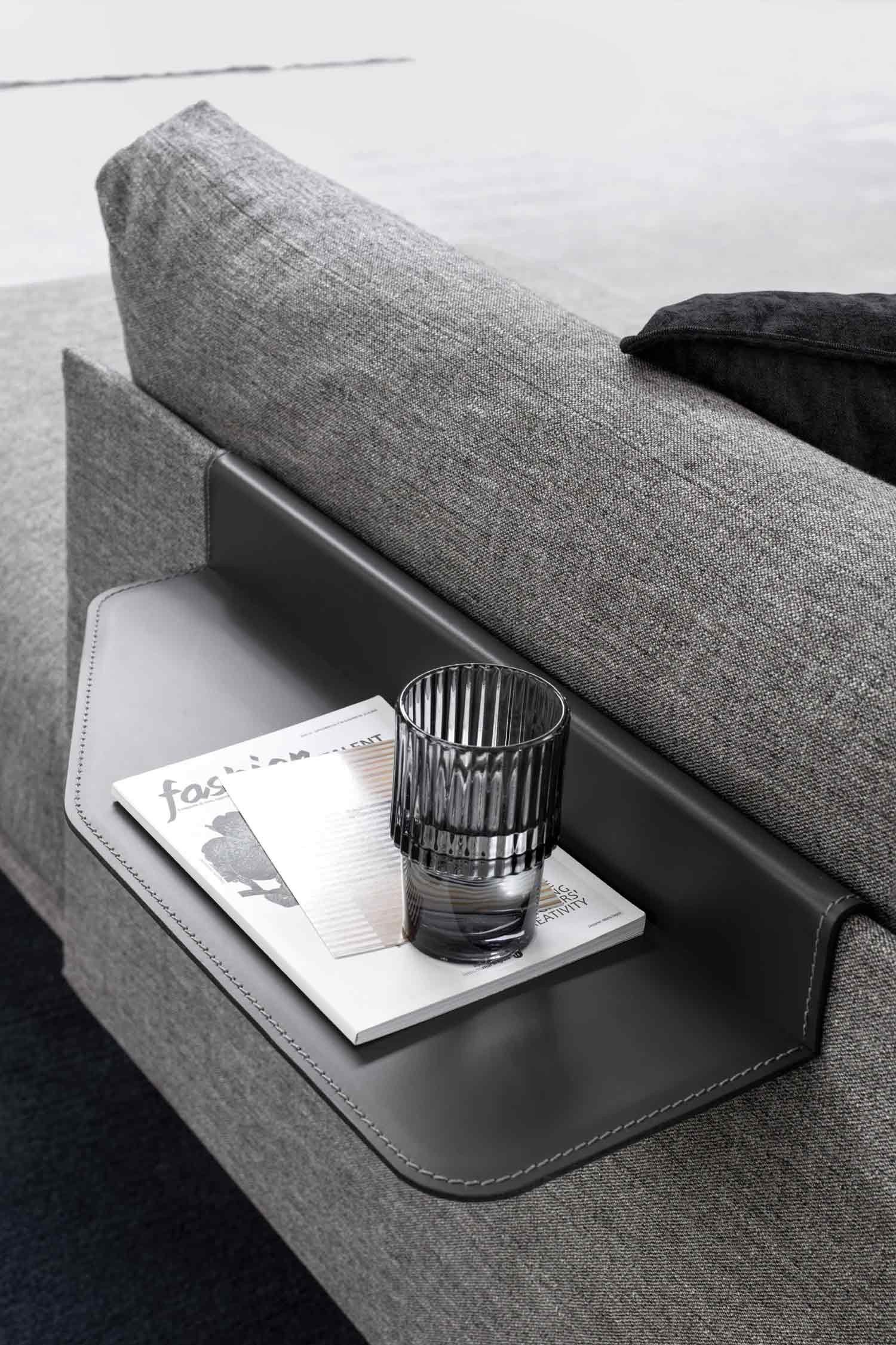 A detailed shot of a black leather armrest tray with white stitching, holding a glass and a magazine on the gray fabric of a sofa.