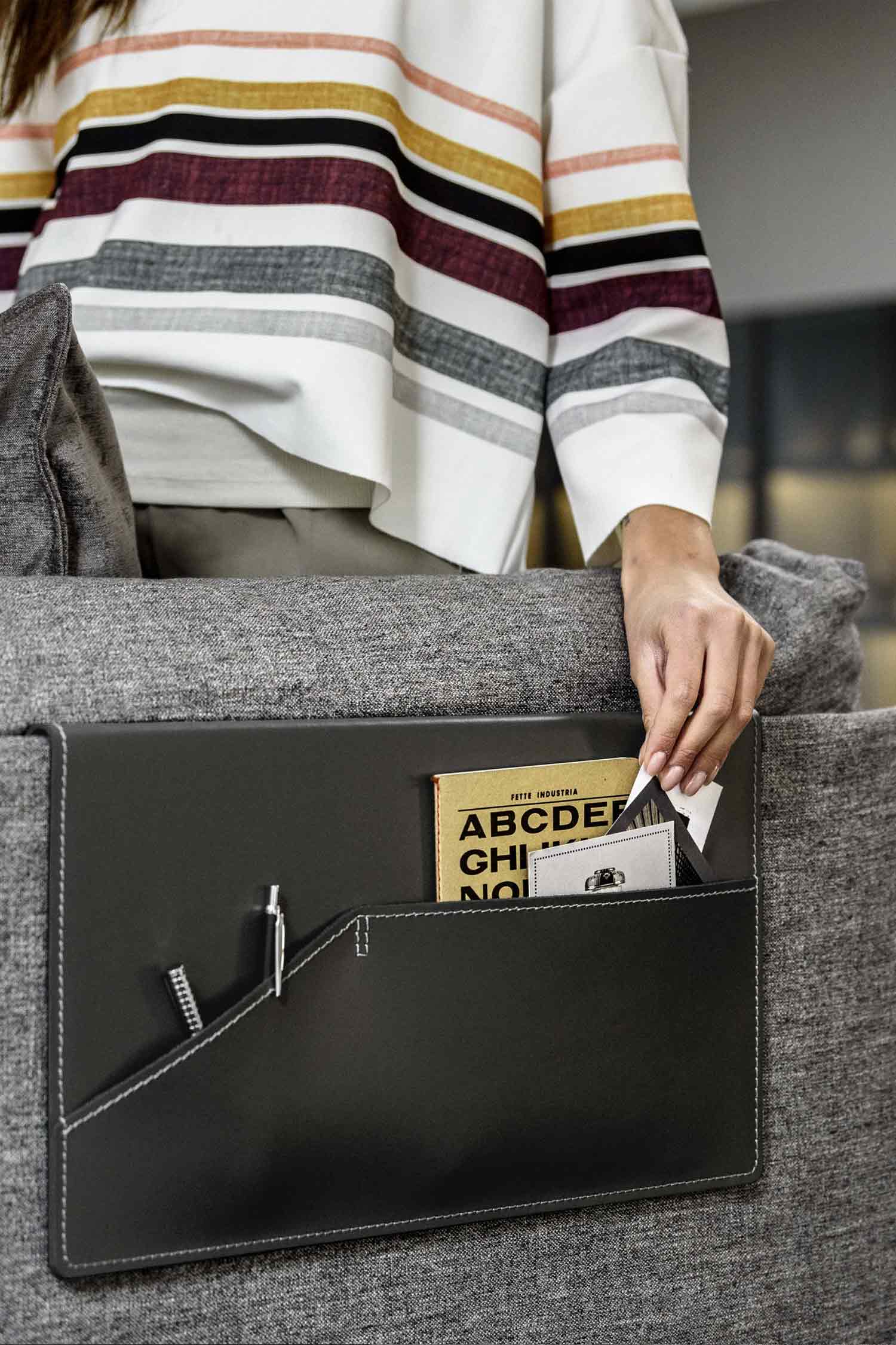 A close-up shot of a person's hand placing an item into a black leather pouch with white stitching, which is attached to the armrest of a gray fabric sofa.
