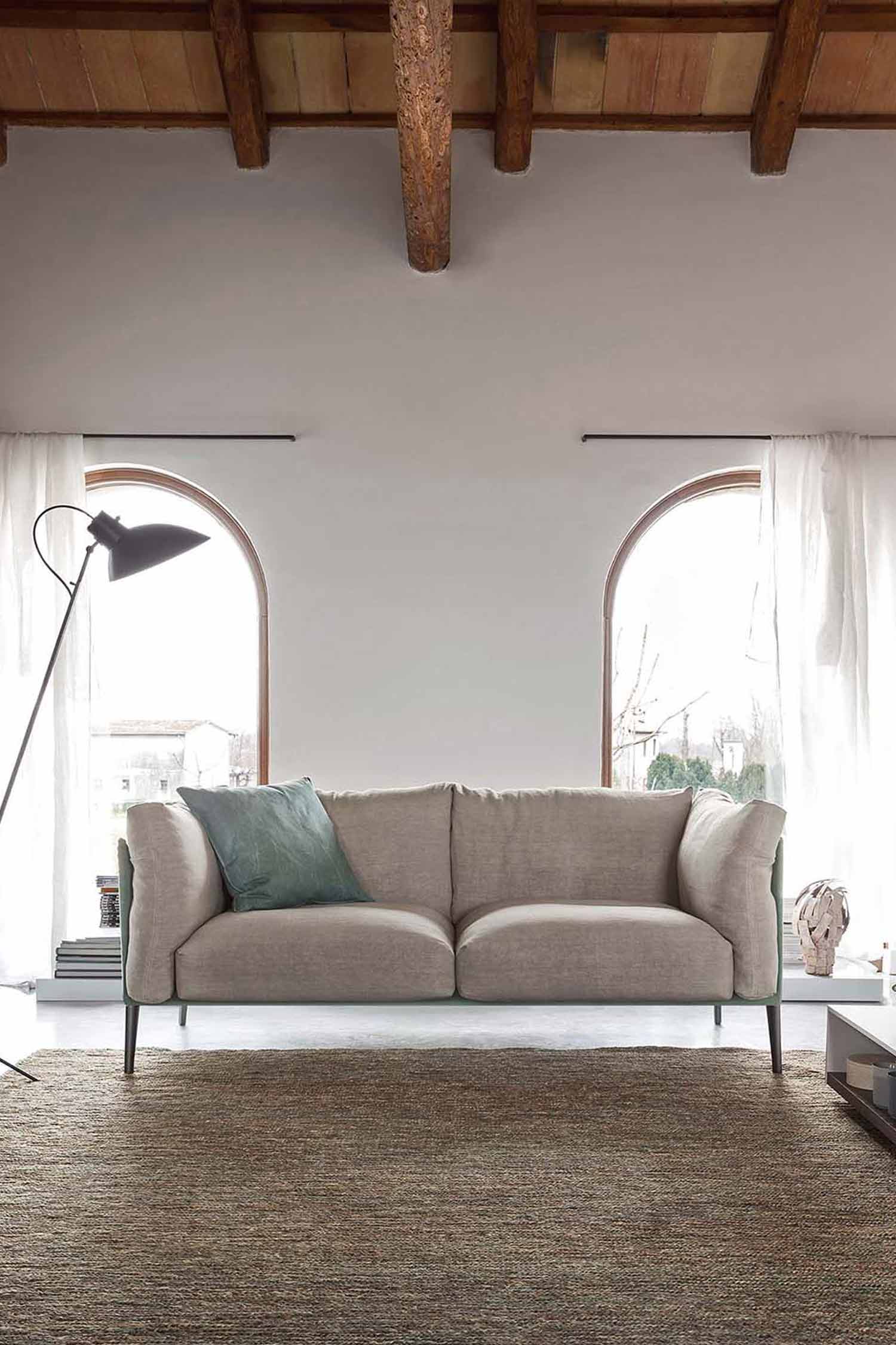 A beige sofa sits on a woven rug in a rustic-style room with exposed wood beams on the ceiling and two tall, arched windows with sheer white curtains.