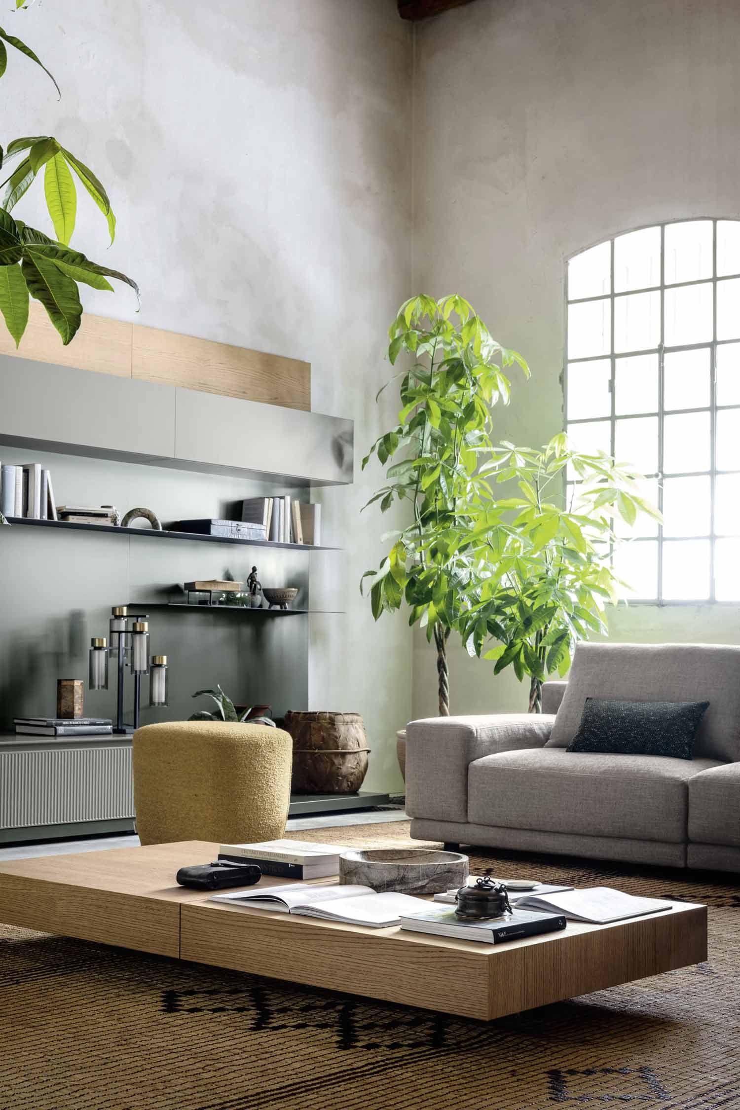 A stylish living room with a large, light wood coffee table in the foreground. A modular light gray sofa with a black accent pillow is visible to the right, next to a tall, leafy green plant. In the background, there is a gray wall unit with floating shelves and a yellow ottoman.
