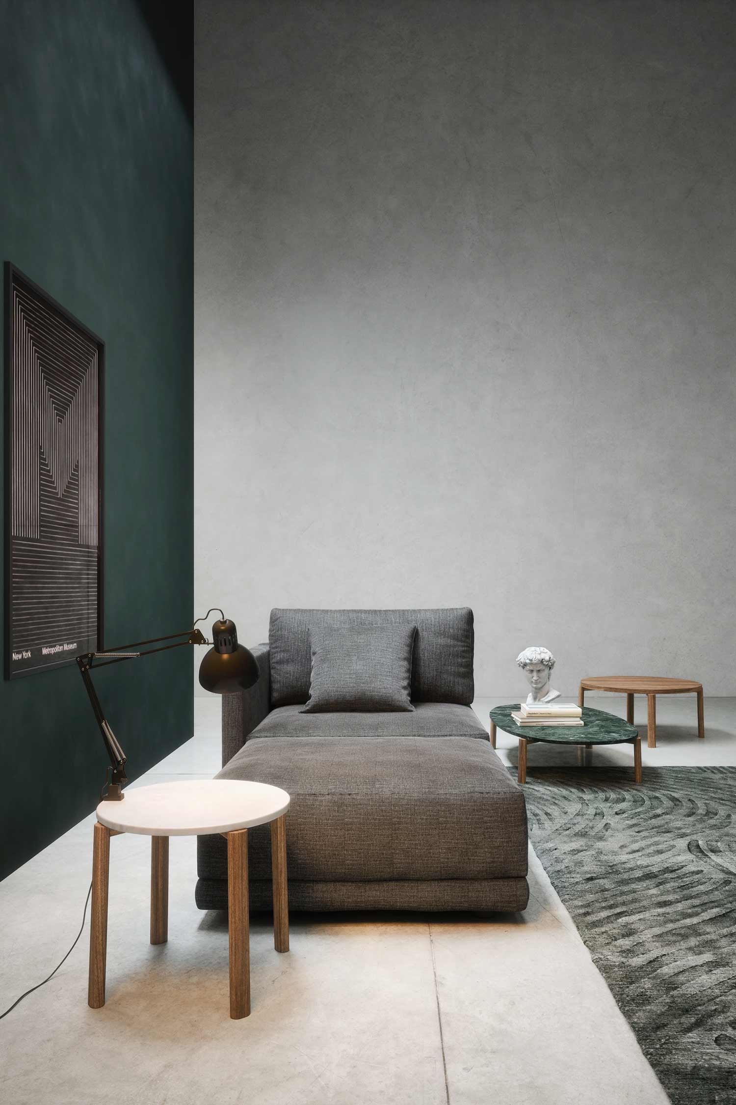 A low-profile gray chaise lounge with a headrest pillow and a single accent pillow. The furniture is set against a dark teal and gray wall. In the foreground, there's a white and wood side table with a black desk lamp, and in the background, a small bust sculpture and three round tables sit on a green and gray textured rug.