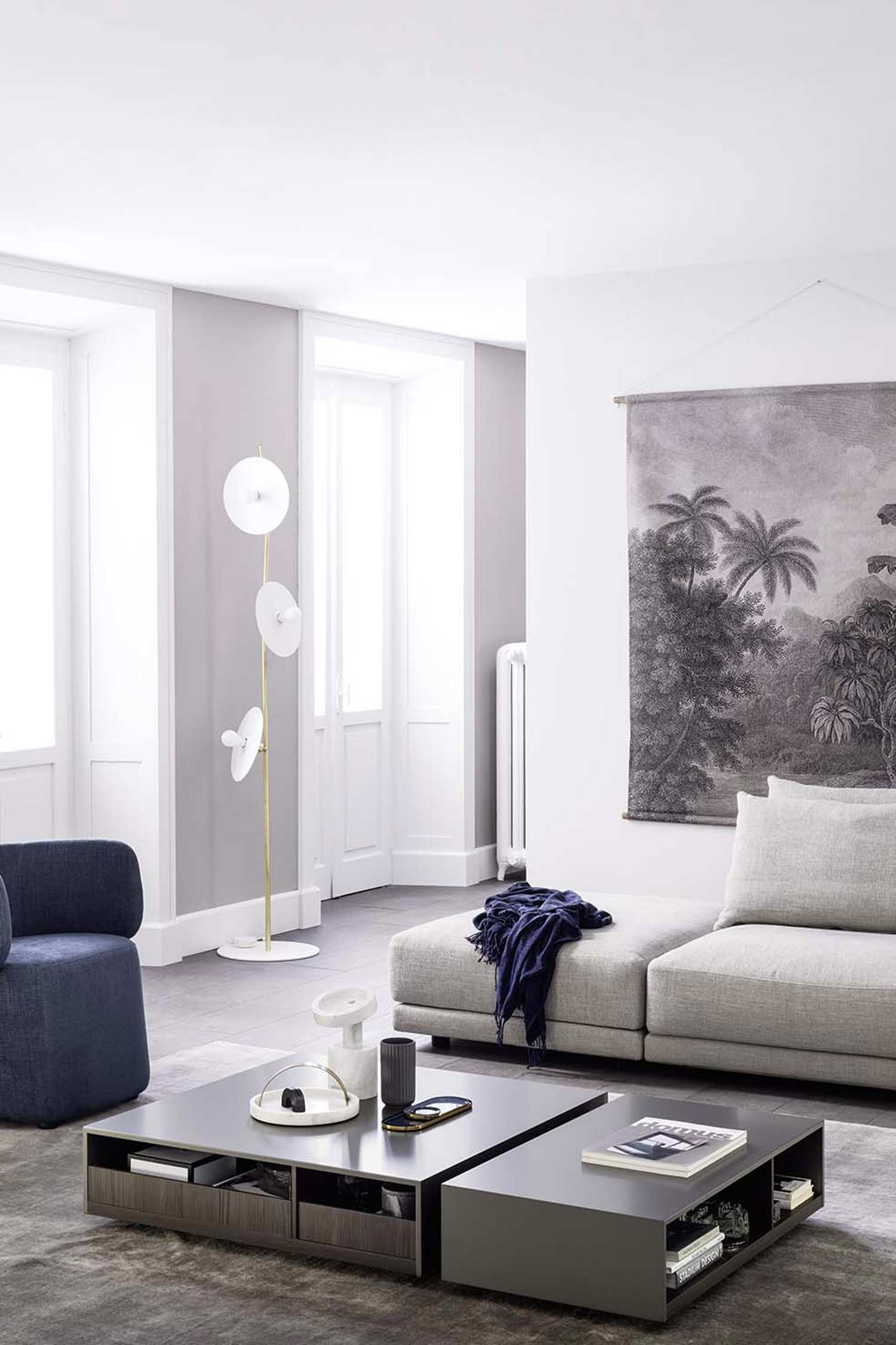 A minimalist living room featuring a gray modular sofa and a blue armchair. A large, two-tiered dark gray coffee table with books and decorative items sits in the center. The room has light gray walls, a tall white floor lamp, and a large, black-and-white tapestry of a jungle scene hanging on the wall.