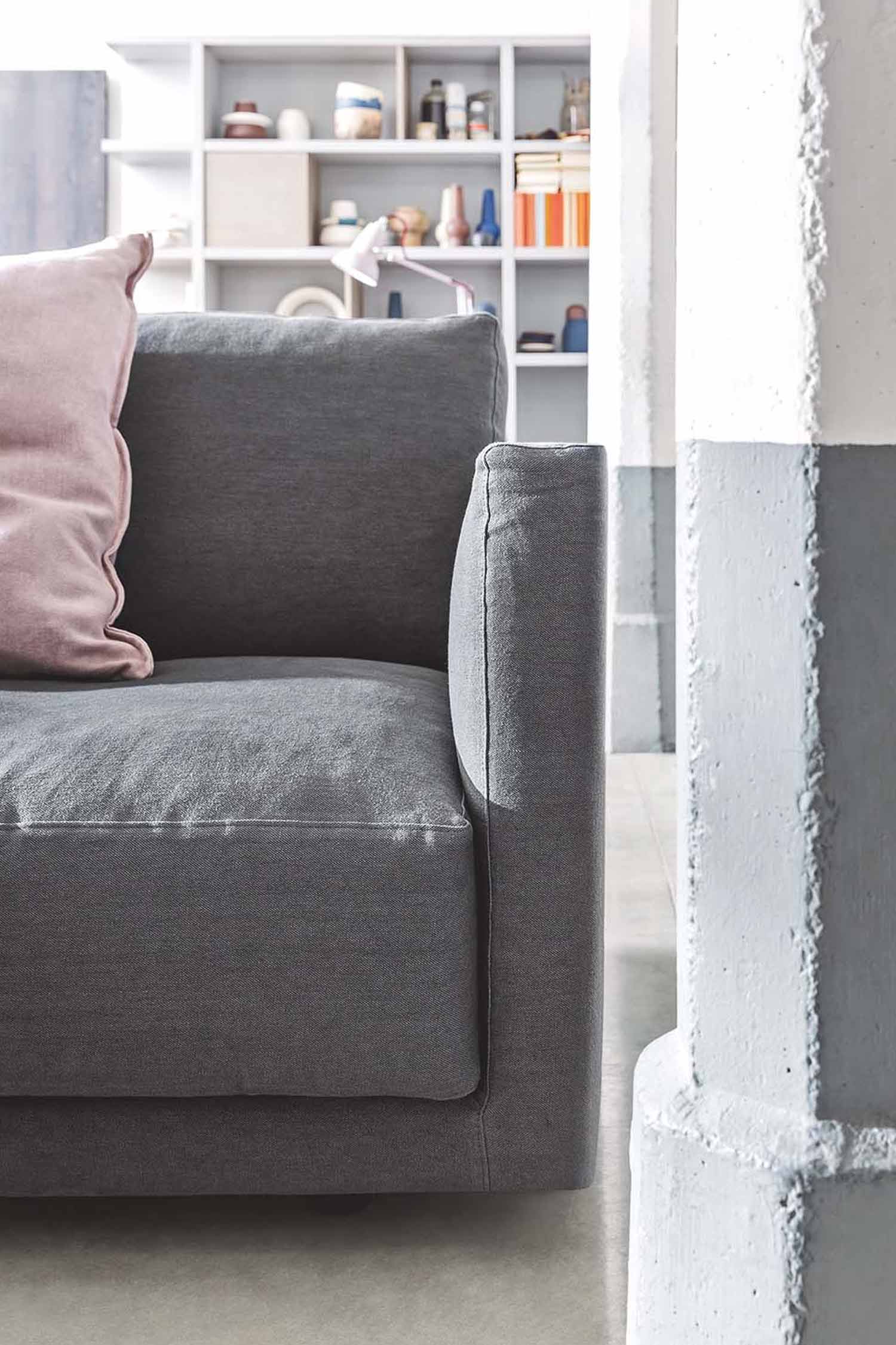 A close-up of a gray modular sofa with a light pink pillow. The side of the sofa is in the foreground, with a rough-textured white and gray pillar to the right and a white bookshelf with colorful objects in the background.