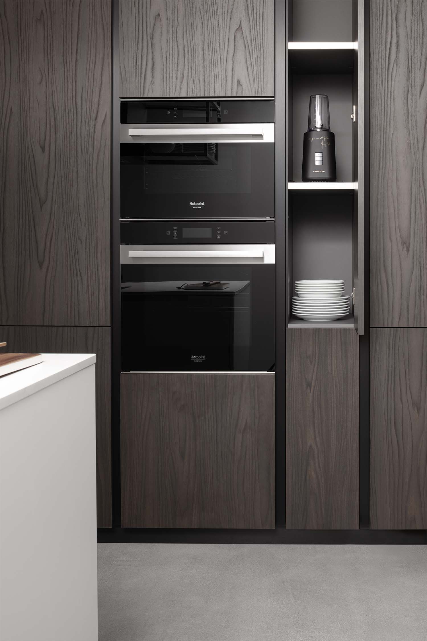 A close-up of a stacked set of black built-in appliances, including a coffee machine, microwave, and oven, housed within dark wood-grain cabinets. An open vertical cabinet to the right reveals a blender and a stack of white plates on a shelf, highlighting the kitchen's integrated storage solutions.