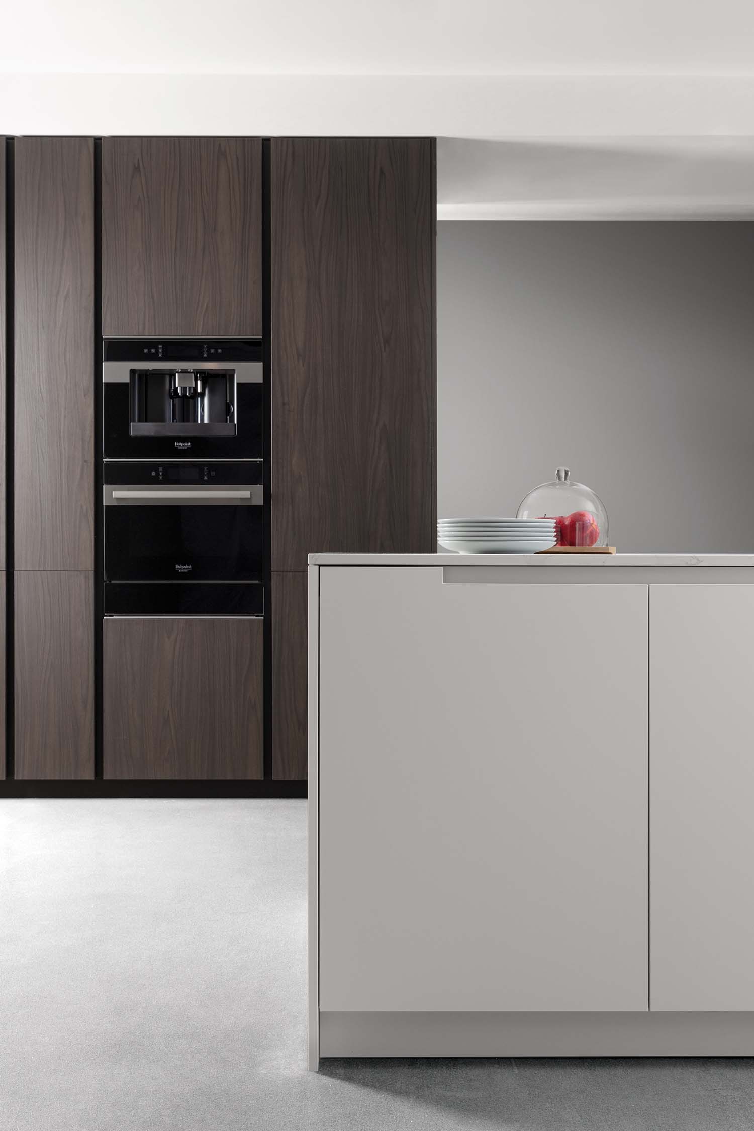 A view of a kitchen island in the foreground and tall cabinets in the background. The island is a simple, white handleless design with a subtle texture and a light, marble-patterned countertop. The dark wood-grain cabinets behind it contain a sleek stack of black built-in appliances.