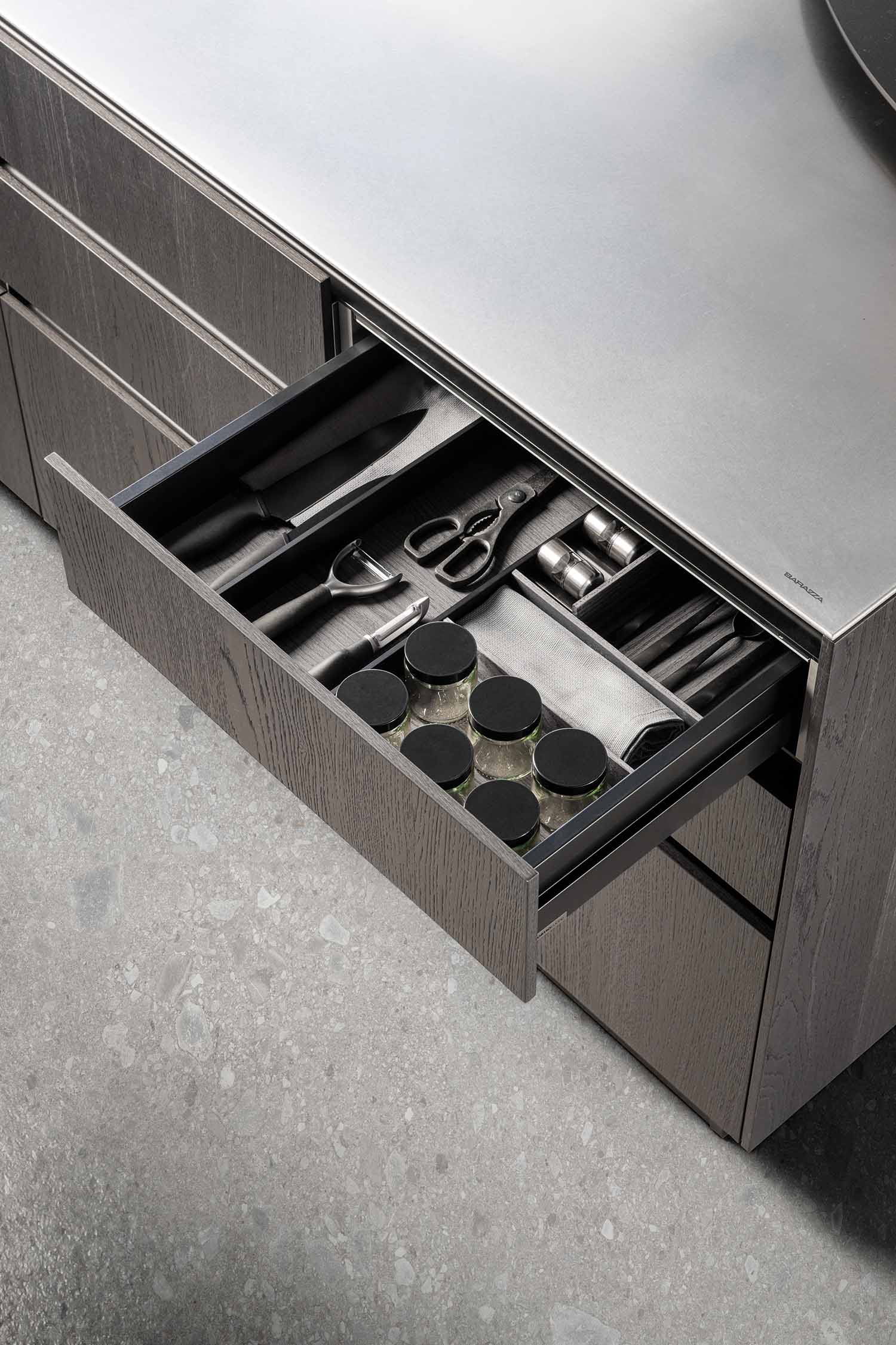A high-angle shot of a kitchen island showing a seamless transition between a dark wood veneer surface and a stainless steel countertop. The stainless steel section includes a built-in hob with a black extractor fan in the center.